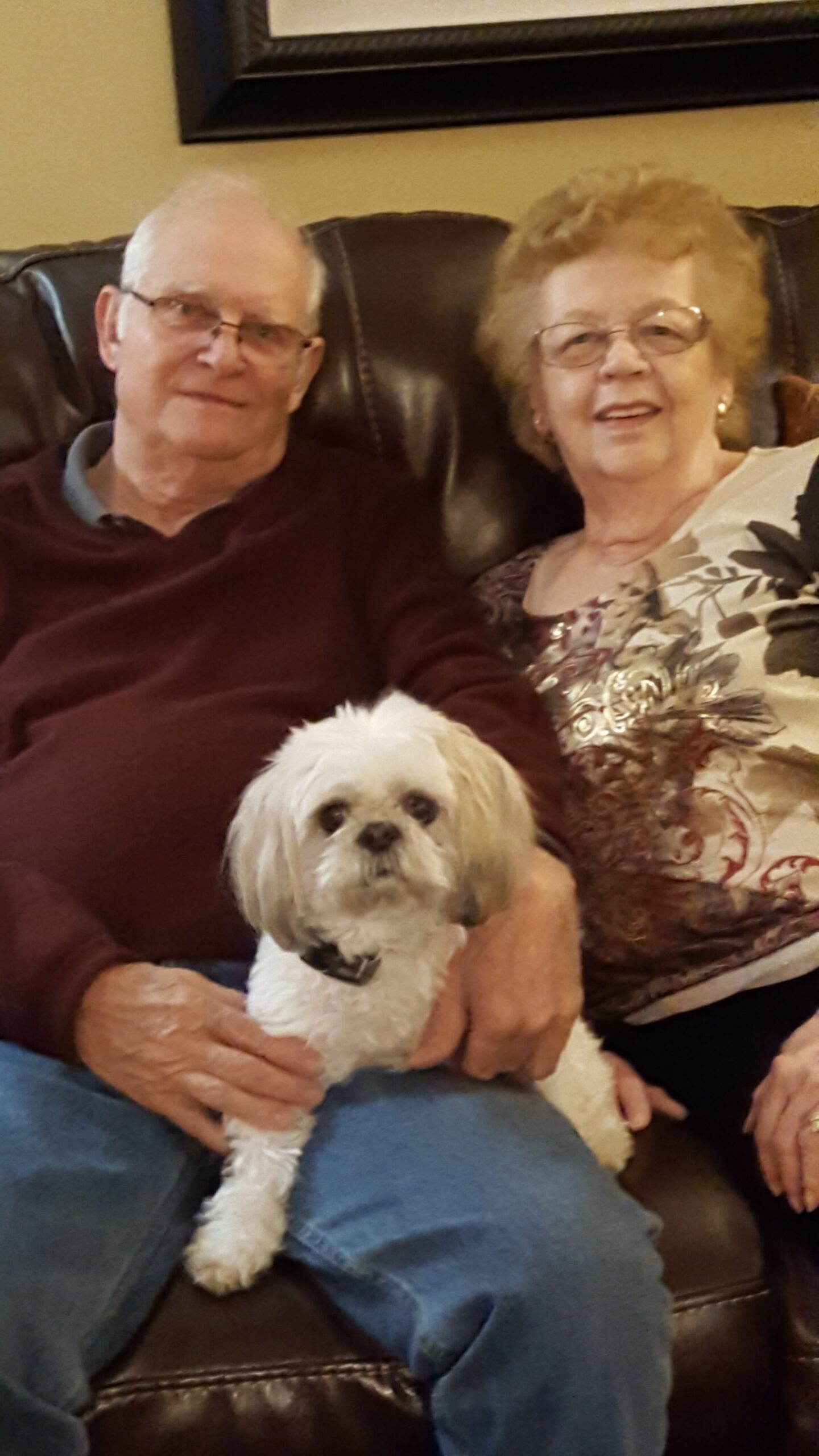 Older couple sitting on a leather couch with a small white dog on the man's lap, smiling at the camera.