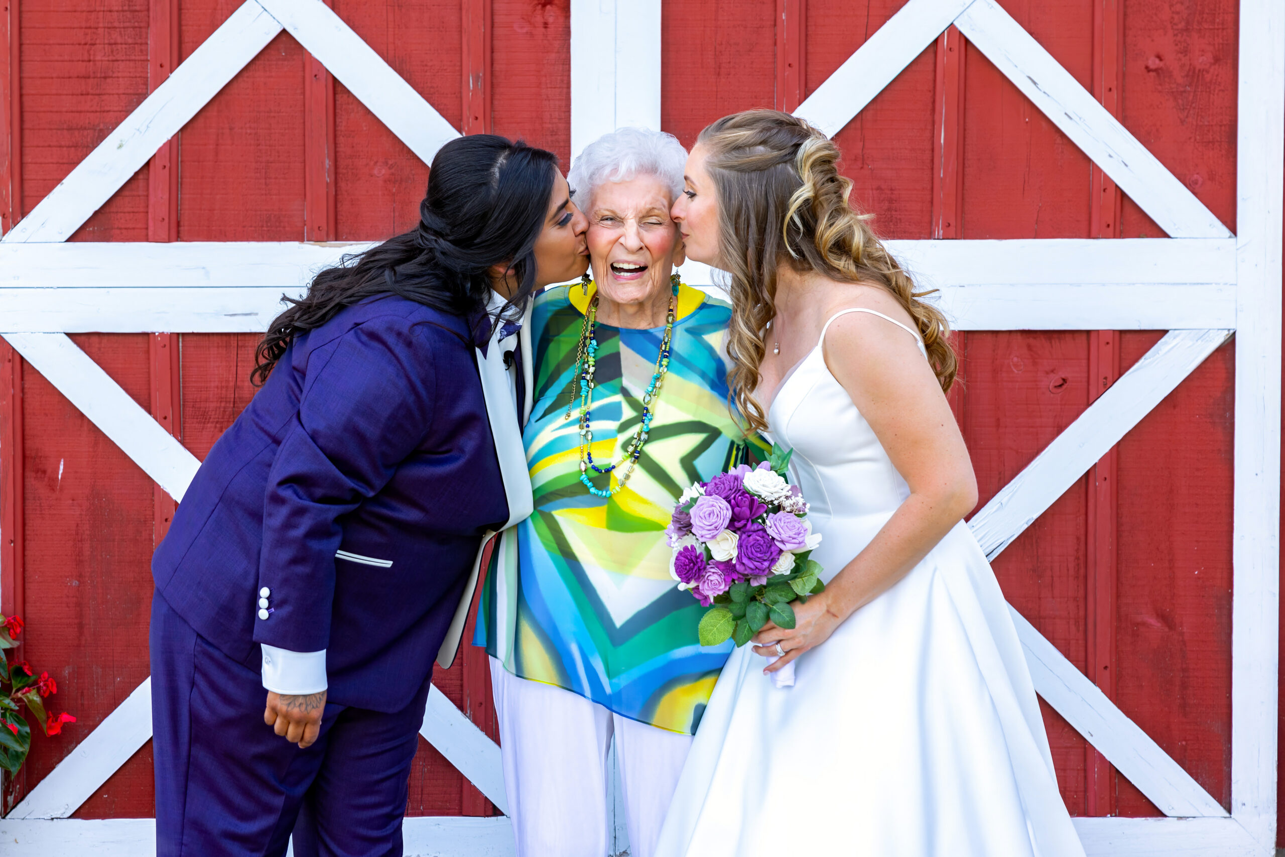 Three generations sharing a kiss on the cheeks: an elderly woman in a bright, patterned dress between two younger women in formal attire, holding a purple bouquet against a red and white barn backdrop.