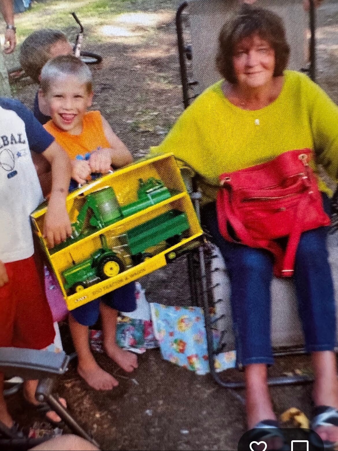 Smiling boy holding a yellow toy tractor box next to a seated woman in a bright yellow shirt and red bag, outdoors.