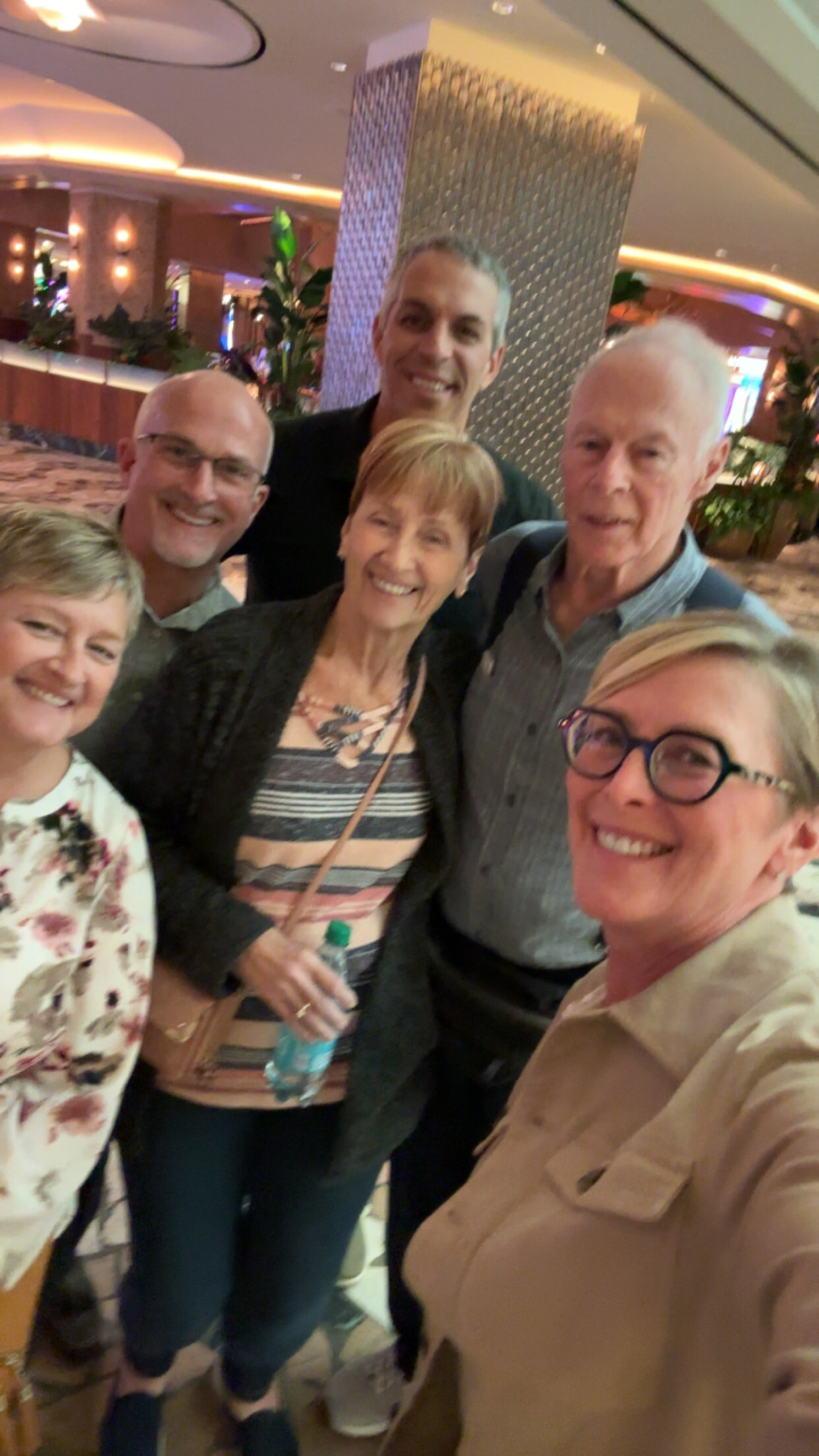 Group of five adults taking a cheerful selfie in a hotel lobby or lounge area, smiling at the camera with warm lighting and plants in the background.
