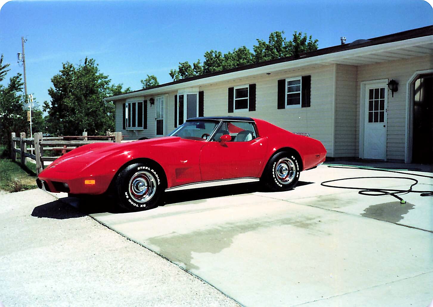 Red vintage Chevrolet Corvette convertible parked on a concrete driveway in front of a beige house with black shutters.
