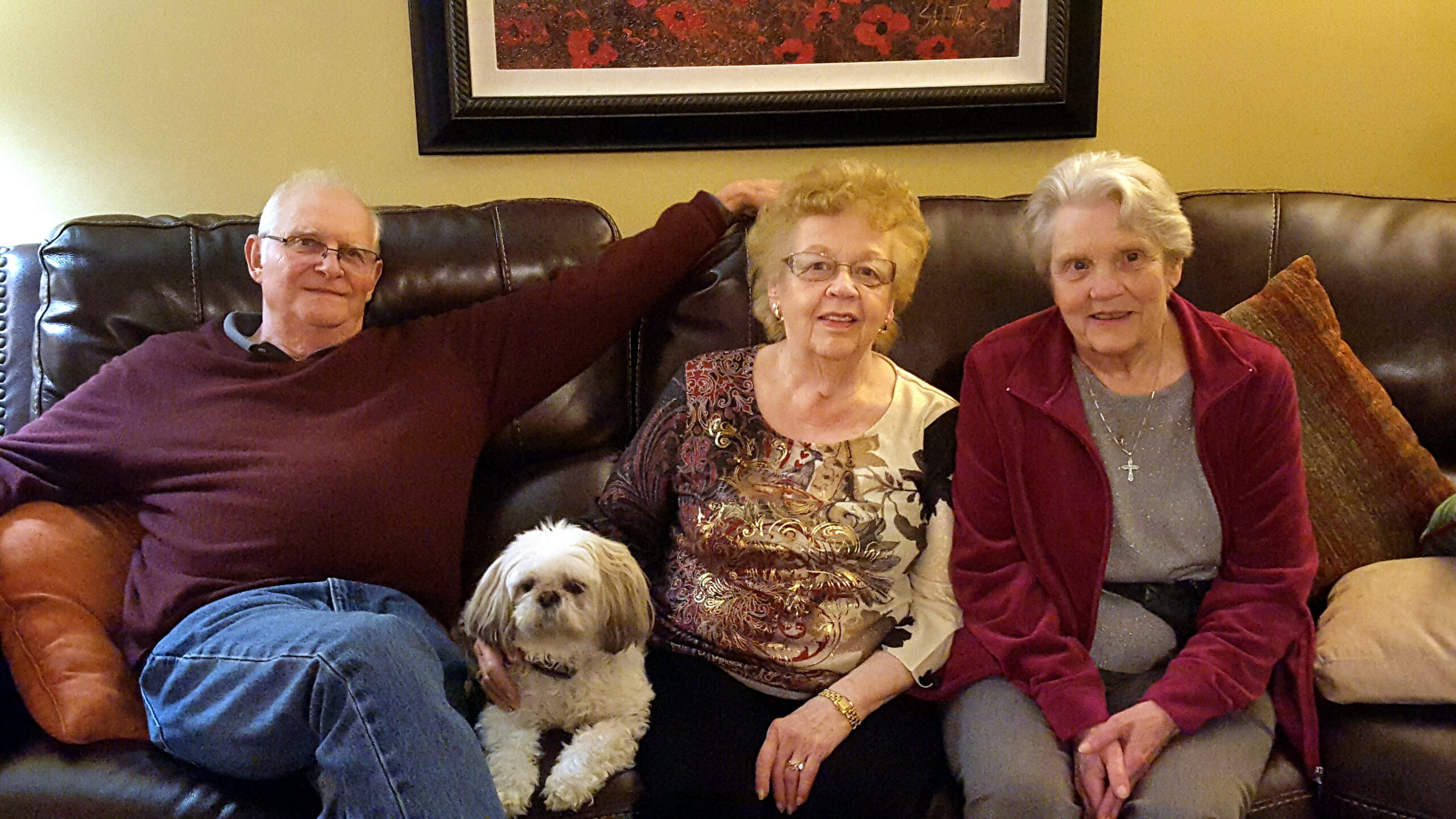 Three seniors seated on a brown leather couch with a small white dog at the center of the group.
