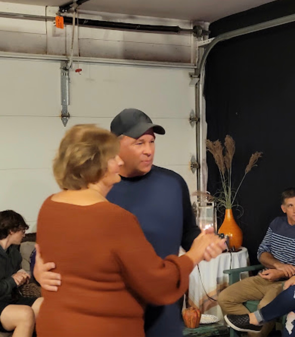 Couple dancing at a casual indoor gathering; woman in brown sweater and man in a dark shirt and cap in the foreground, others seated in the background.