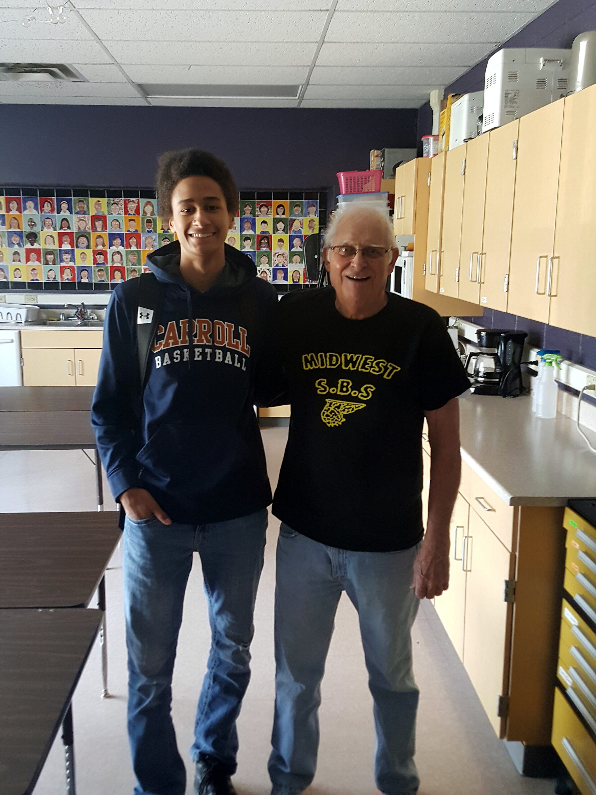 Two men standing together in a school kitchen area, smiling, with wooden cabinets and a coffee station behind them in a classroom setting.
