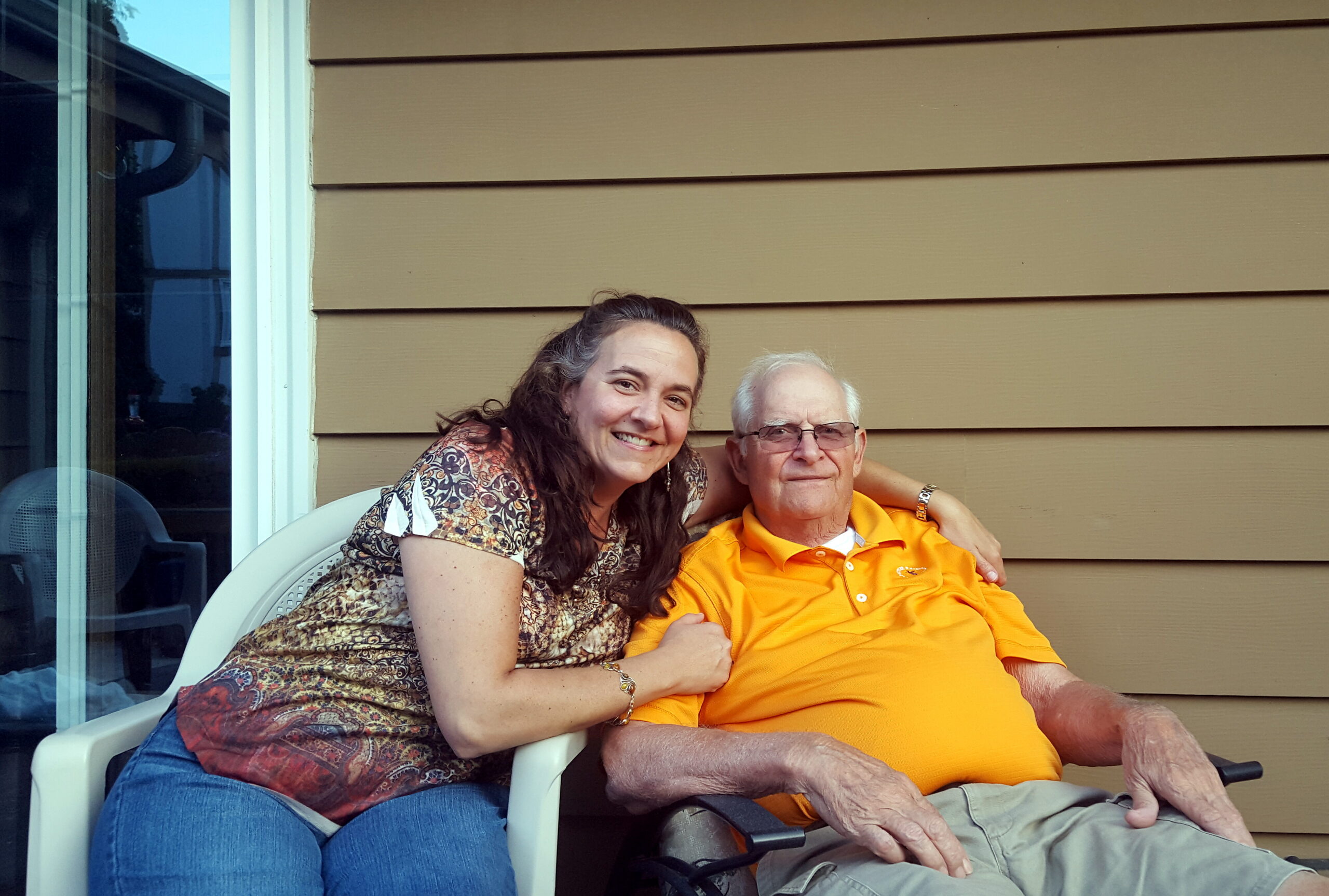 Smiling woman with long hair sitting beside an elderly man in an orange polo shirt on a porch with beige siding behind them, arms around each other.