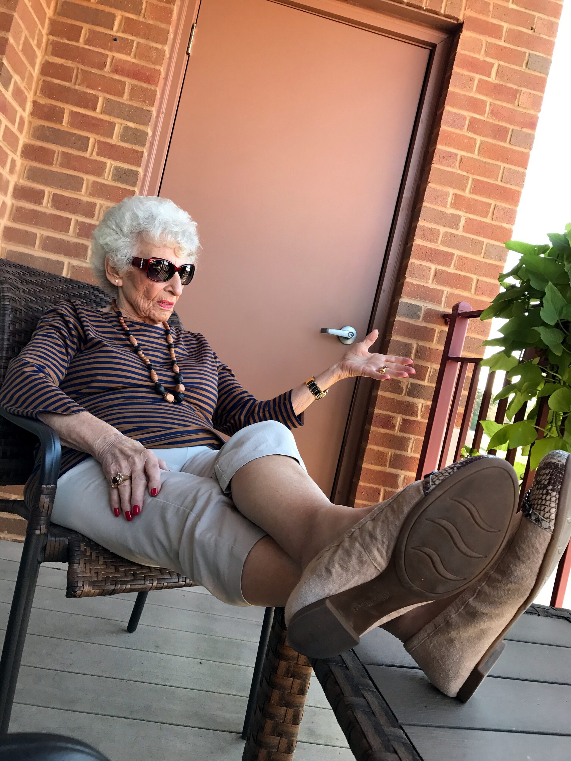 Older woman with white hair wearing sunglasses sits in a wicker chair on a brick porch, gesturing with her hand by a door.