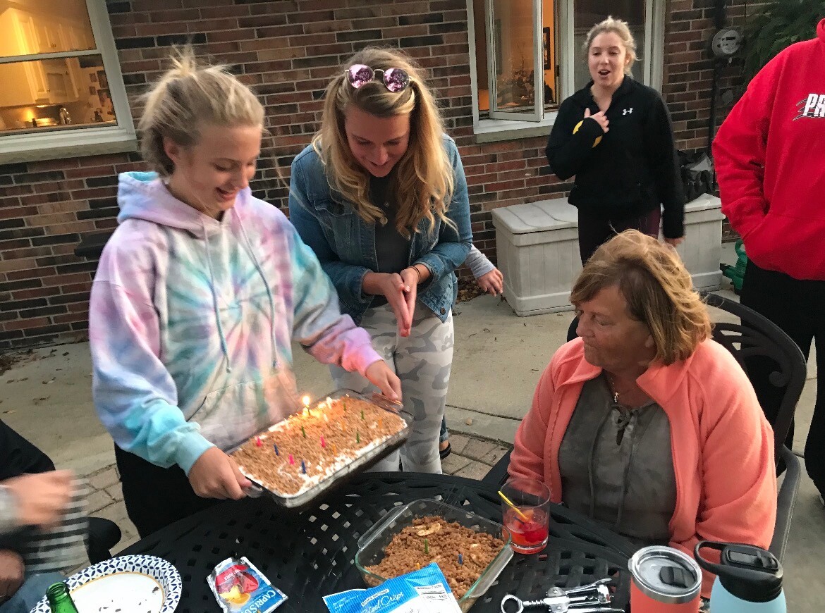 Teenager in a tie-dye hoodie carries a frosted rectangular birthday cake with lit candles toward a seated woman outside on a patio, as friends gather nearby.