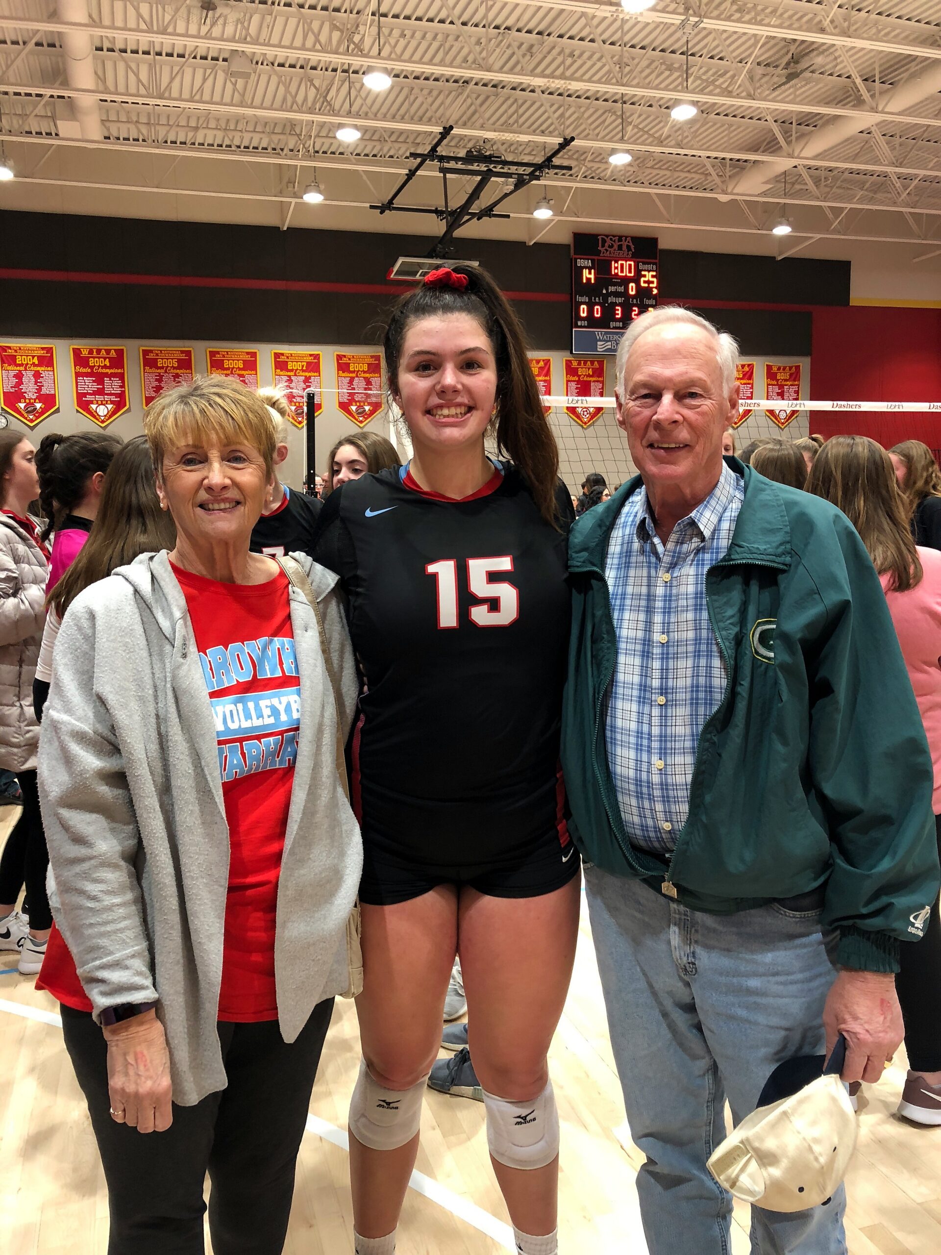Three people pose in a gym after a volleyball event: a smiling player wearing black jersey number 15 with knee pads, flanked by a woman in red and a man in a green jacket, with banners and a scoreboard in the background.