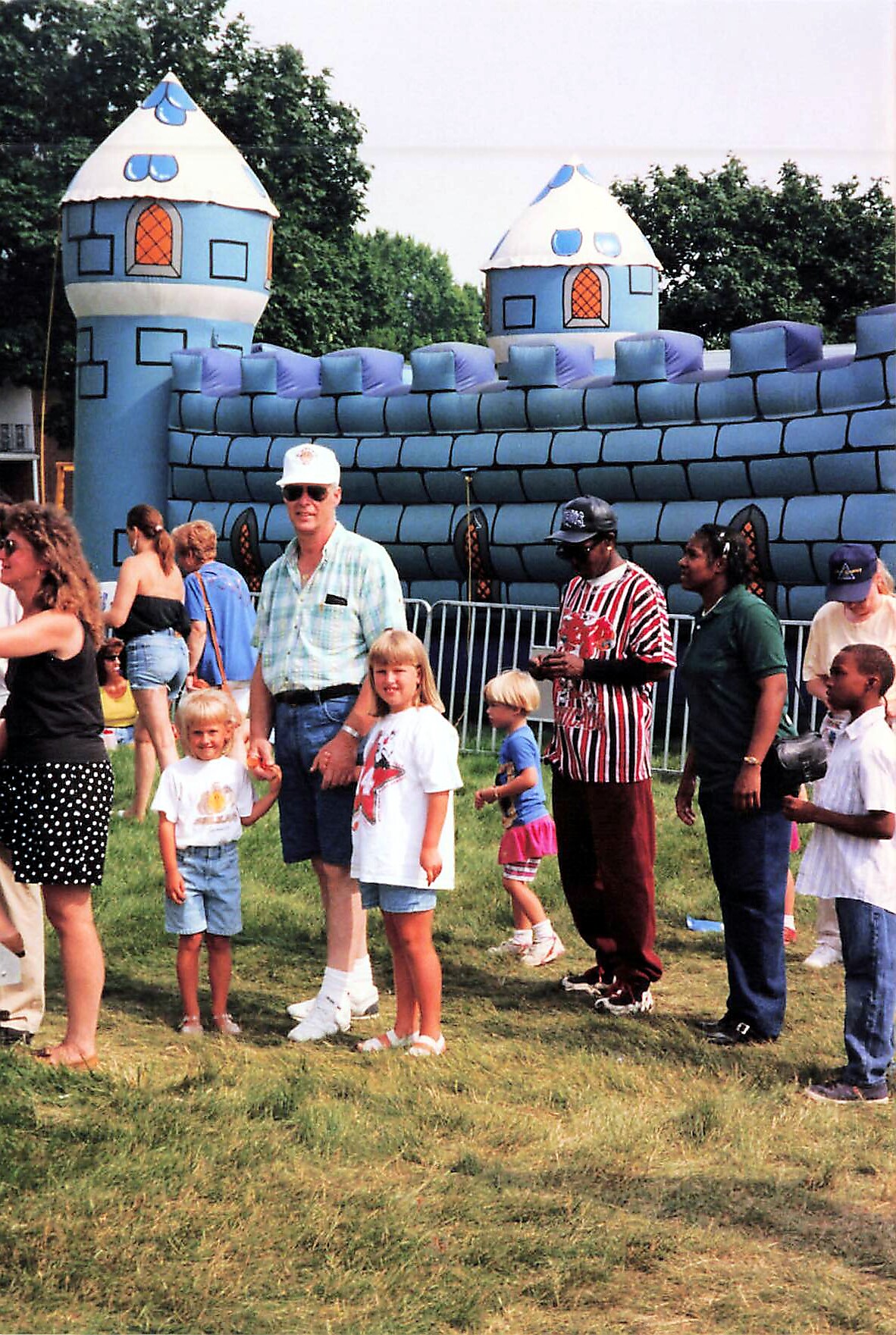 Family-friendly outdoor festival with kids and adults standing on grass near a large blue inflatable castle structure in the background.