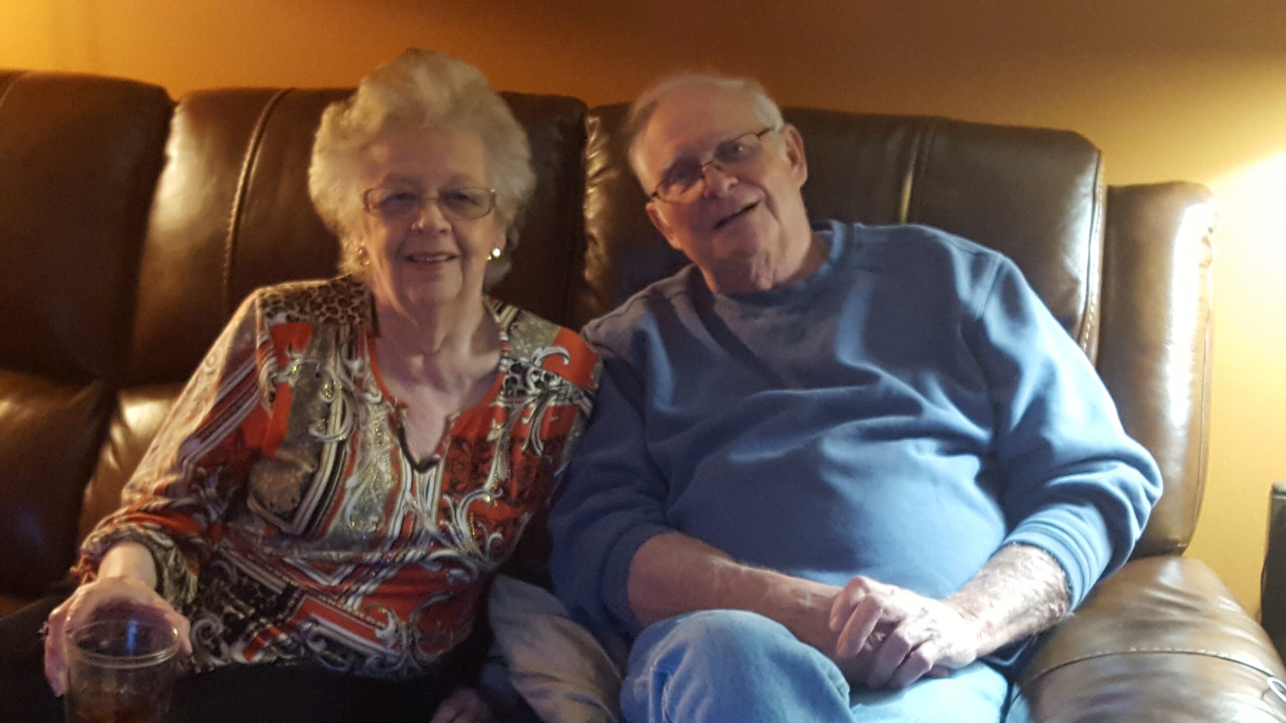 Smiling elderly couple seated on a brown leather couch, the woman holding a glass in her hand.