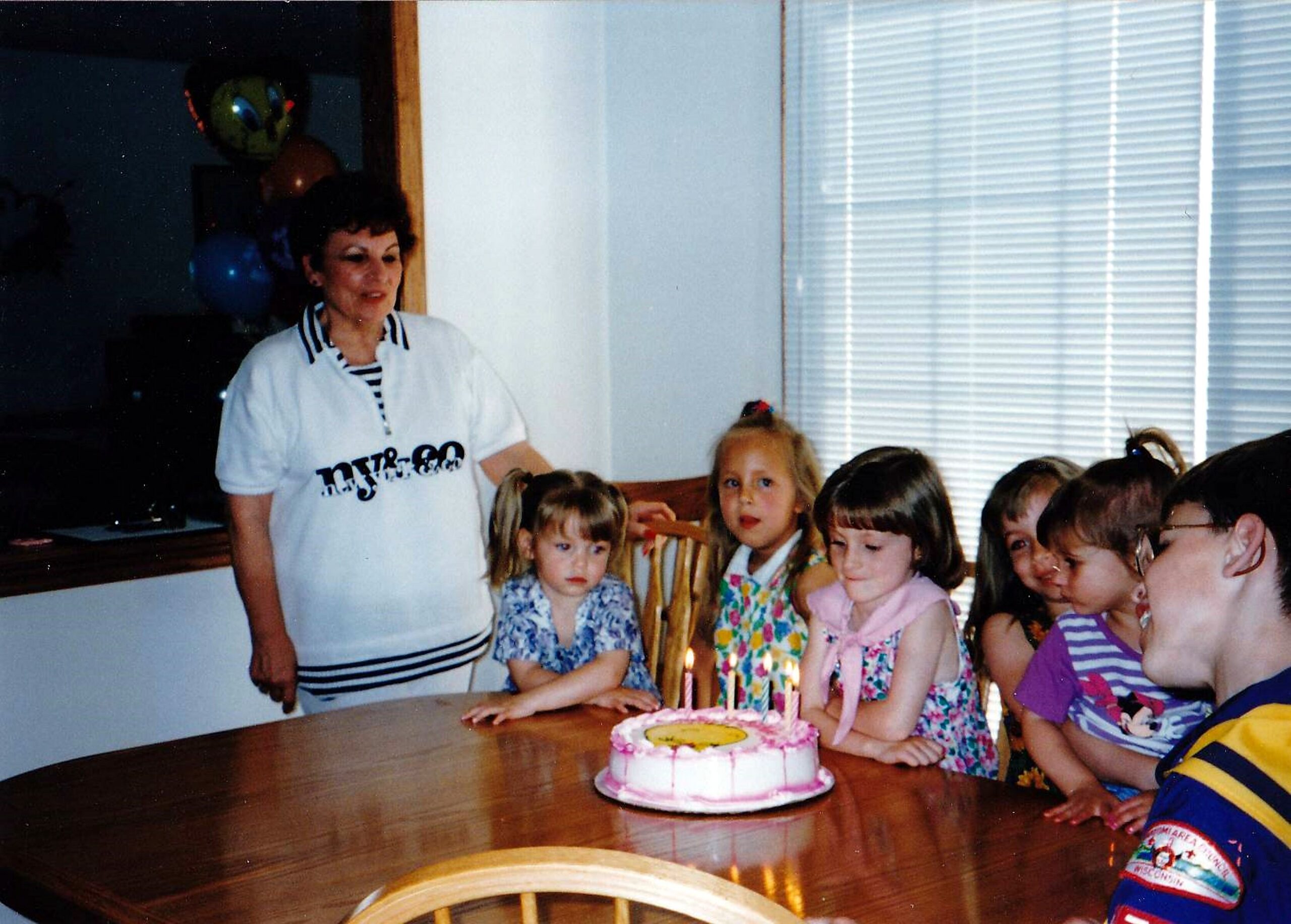 Woman standing beside a table with a pink birthday cake and lit candles, while children gather for a party.