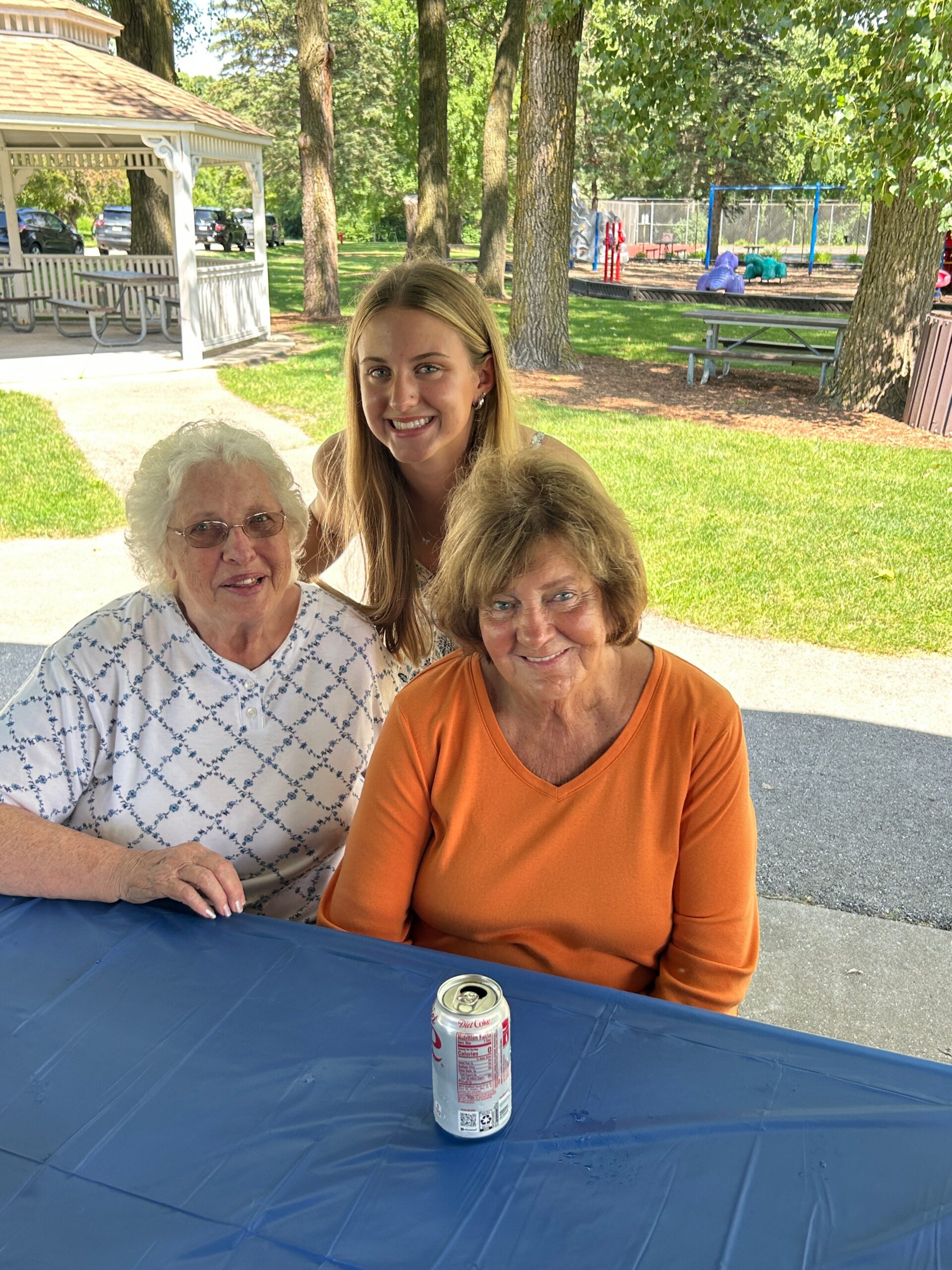 Three smiling women sit at a blue picnic table in a park, with a Diet Coke can in the foreground and a playground in the background.