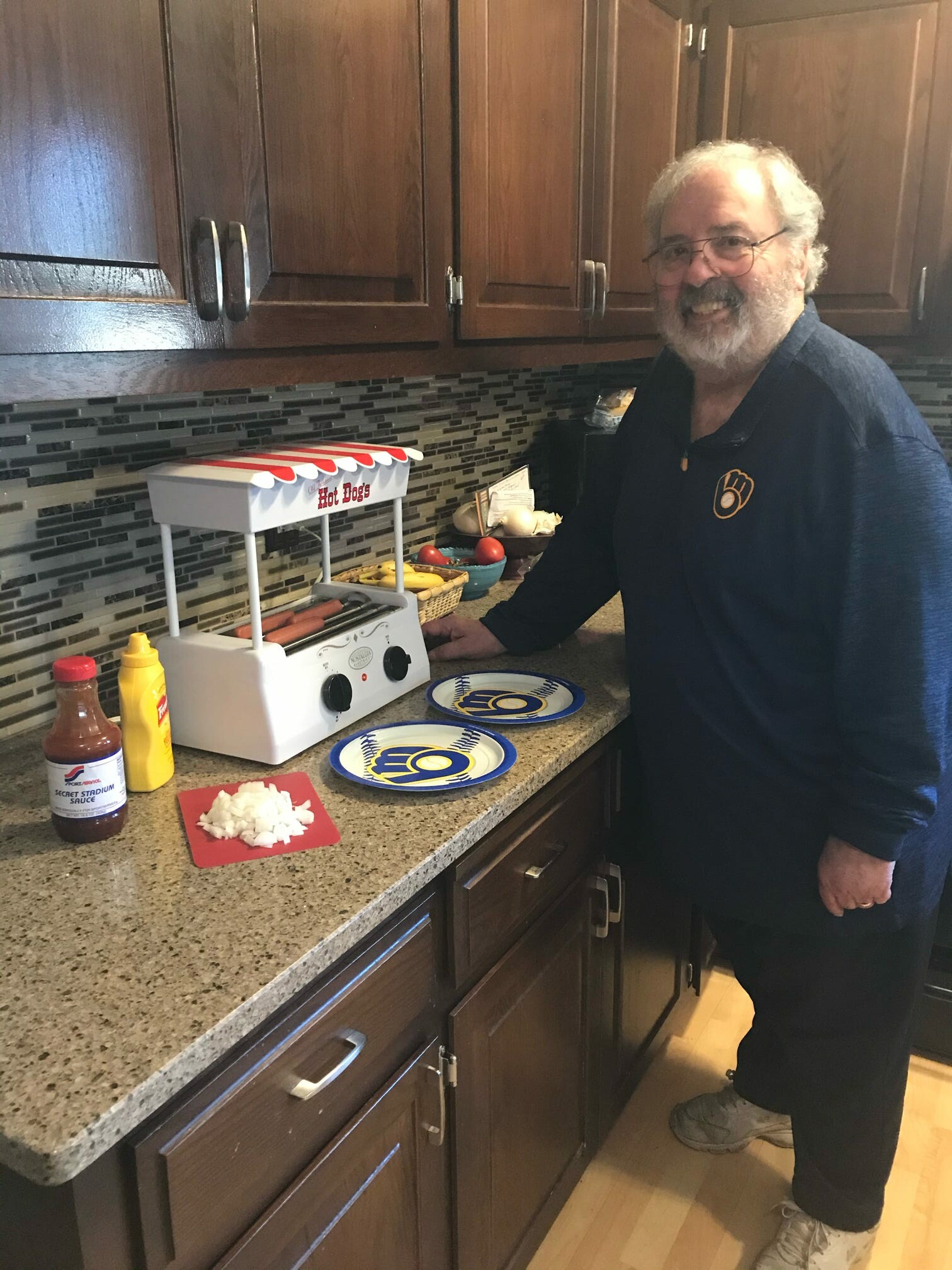 Man in a kitchen using a hot dog toaster, with buns, toppings, and condiments on the counter; preparing hot dogs.