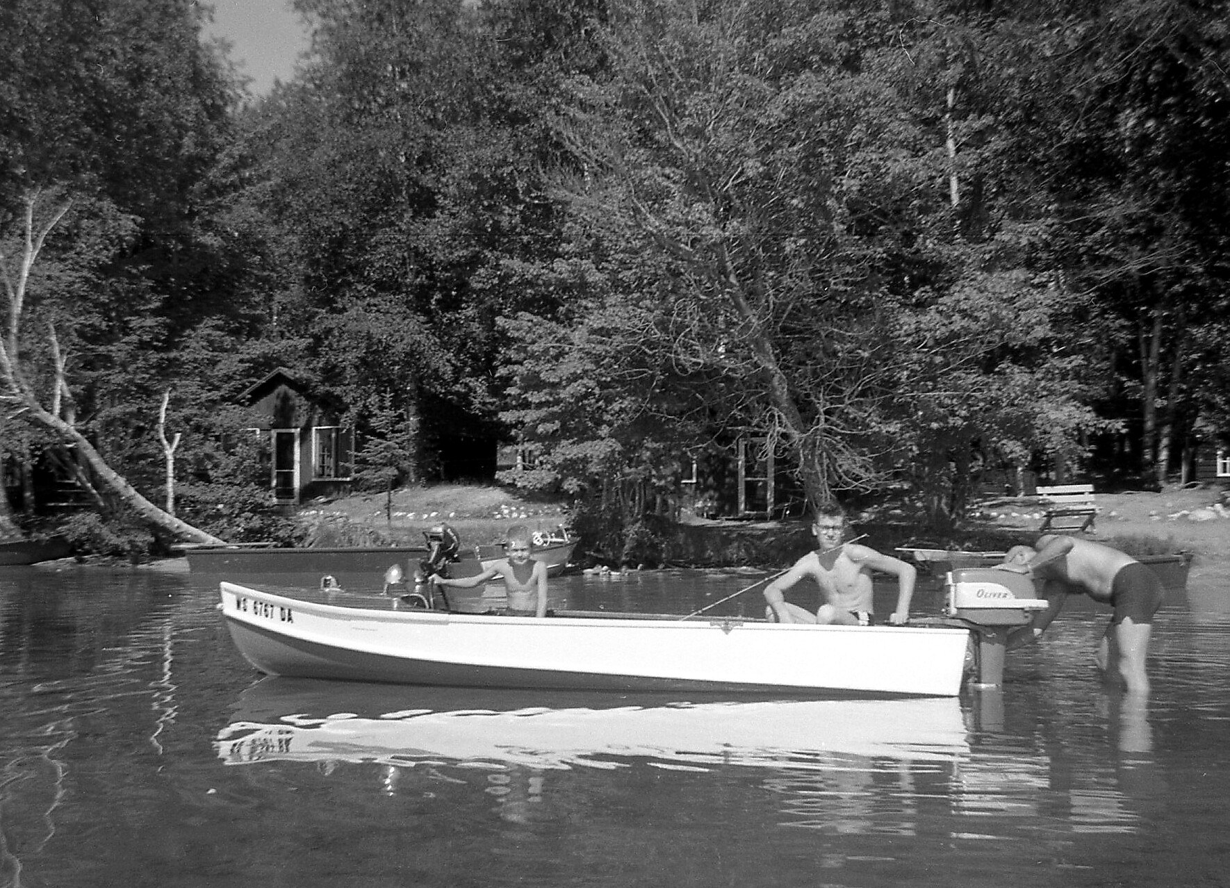 Black-and-white photo of men in a boat on a calm lake, with a wooded shoreline and small boathouse in the background.