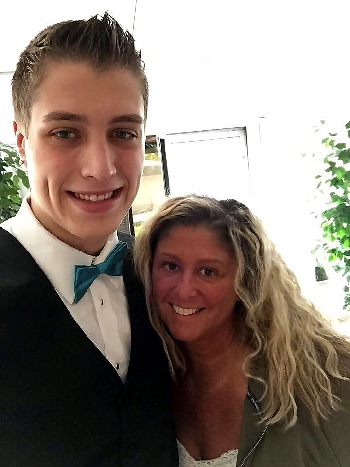 Smiling young man in a black suit and teal bowtie posing with a smiling woman indoors near plants, likely at a formal event.