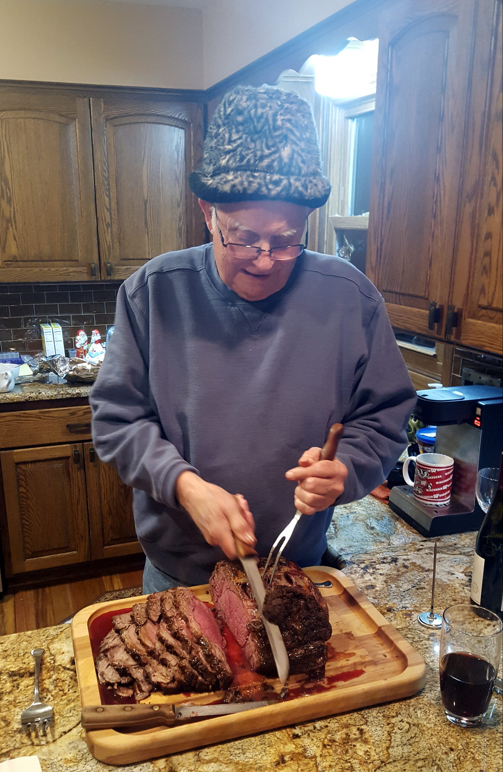 Older man wearing a furry hat slices cooked roast beef on a wooden cutting board in a kitchen.