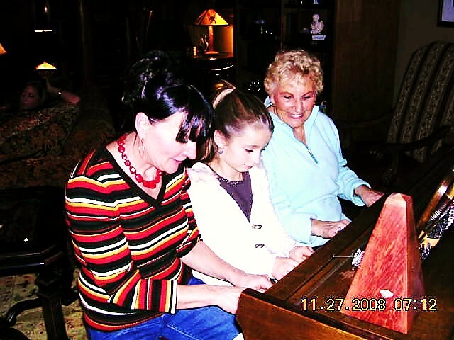 Three people gathered around a wooden piano, with a child between two adults, smiling as they play together.