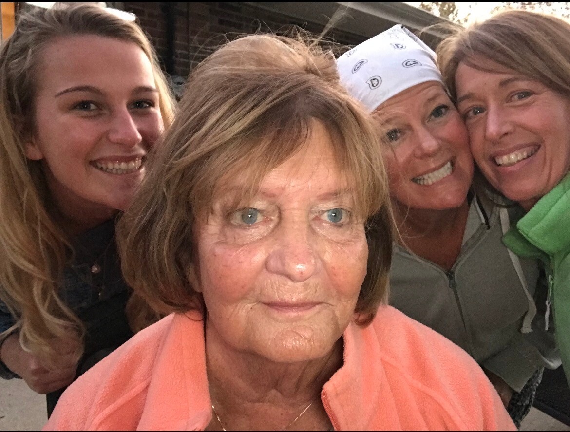 Four women pose for a selfie, with an older woman in a peach jacket at the center smiling broadly.