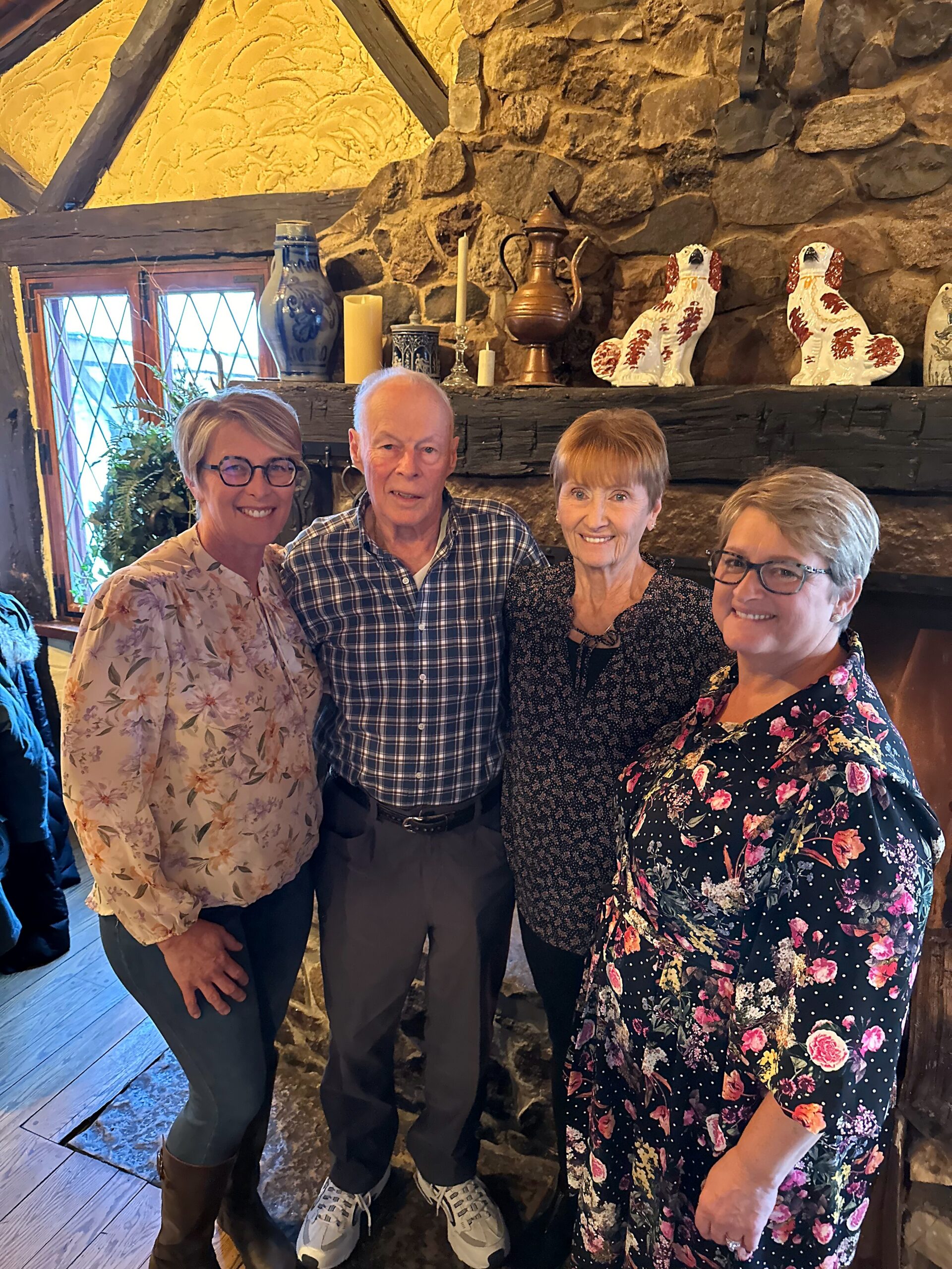 Four adults standing together indoors, smiling, in a rustic stone room with a wooden mantel and decorative ceramic dogs on display.