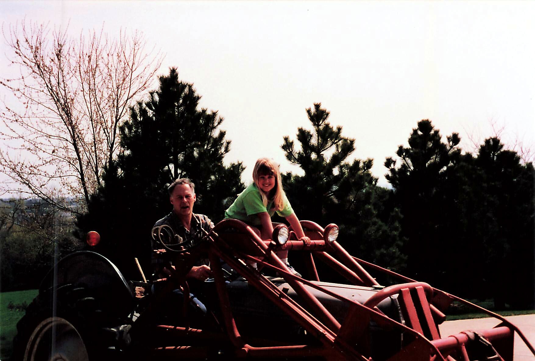 Man and a girl ride a red tractor-like vehicle outdoors, smiling with trees in the background.