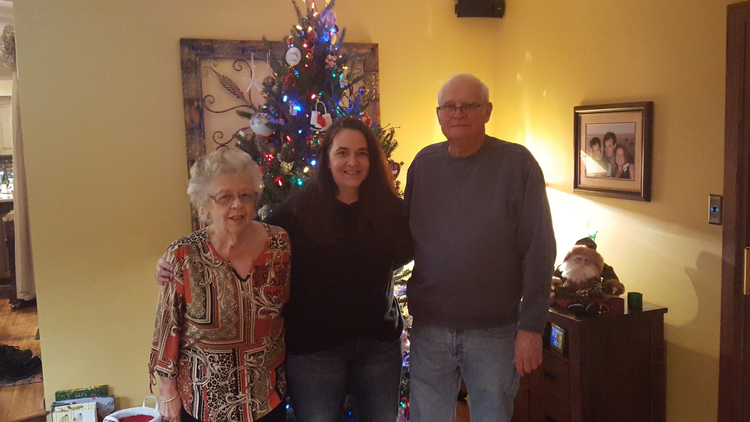 Three people posing indoors in front of a lit Christmas tree with gifts beneath. Left is an elderly woman in a patterned blouse, center is a young woman, right is an older man in a gray sweater.