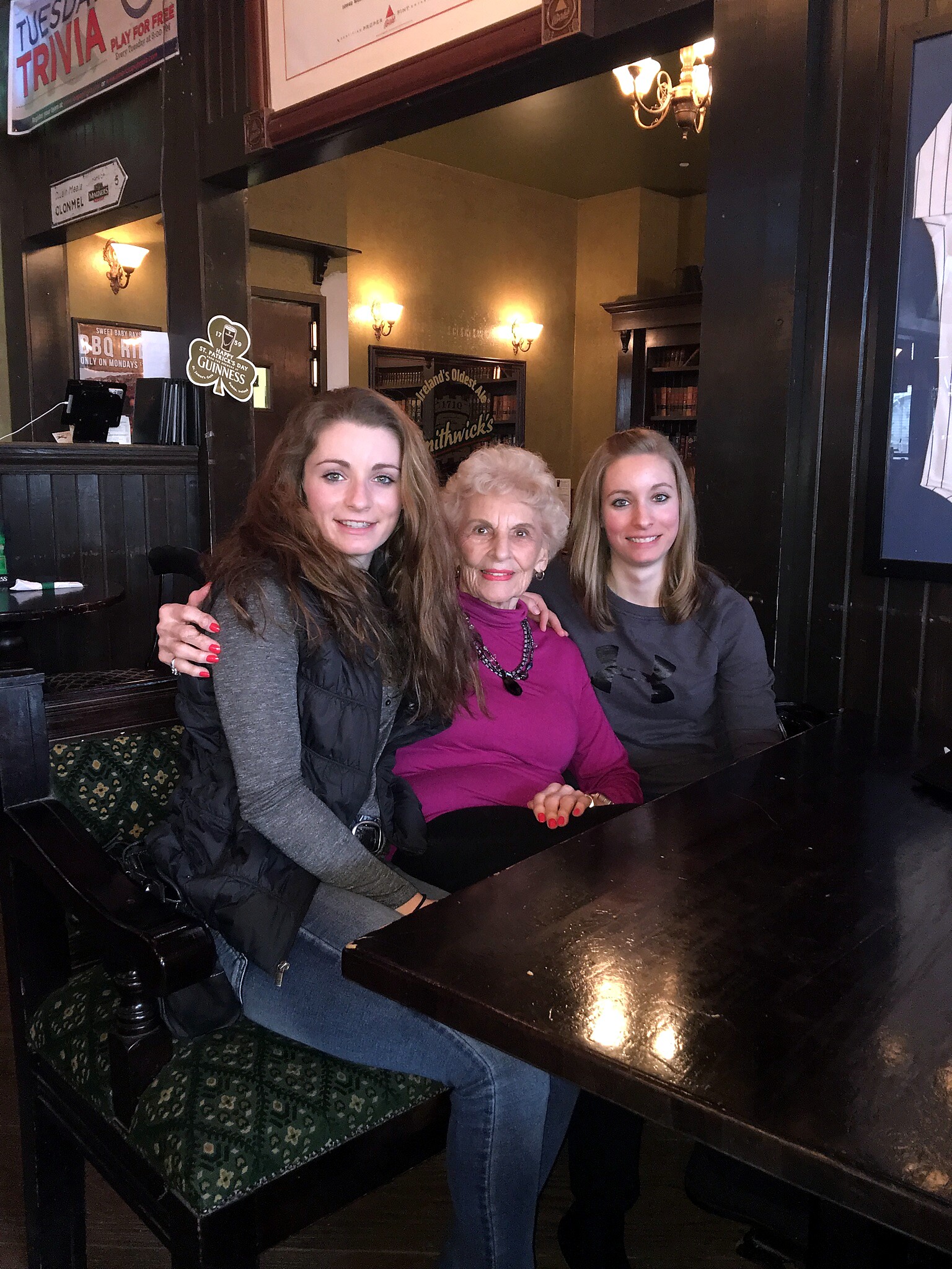Three women posing together at a dark wooden pub table, smiling at the camera; an elderly woman in a magenta top sits between two younger women in gray and black jackets.