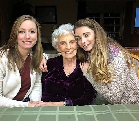 Three women of different ages sit closely together at a table, smiling at the camera indoors, with a green checkered tablecloth in the foreground.