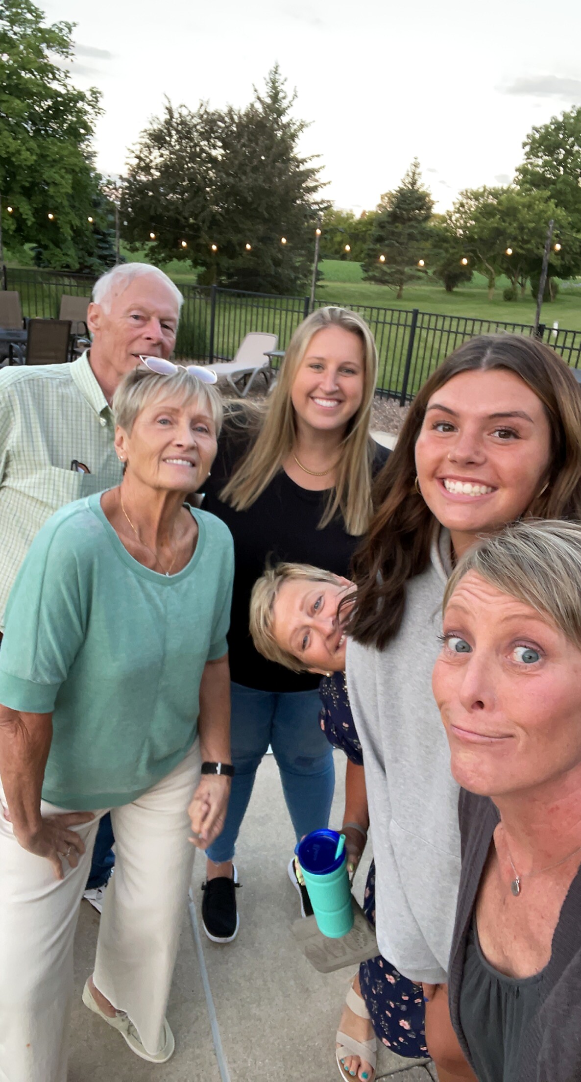 Six friends and family posing for a group selfie outdoors on a patio, string lights, green park in background, everyone smiling.