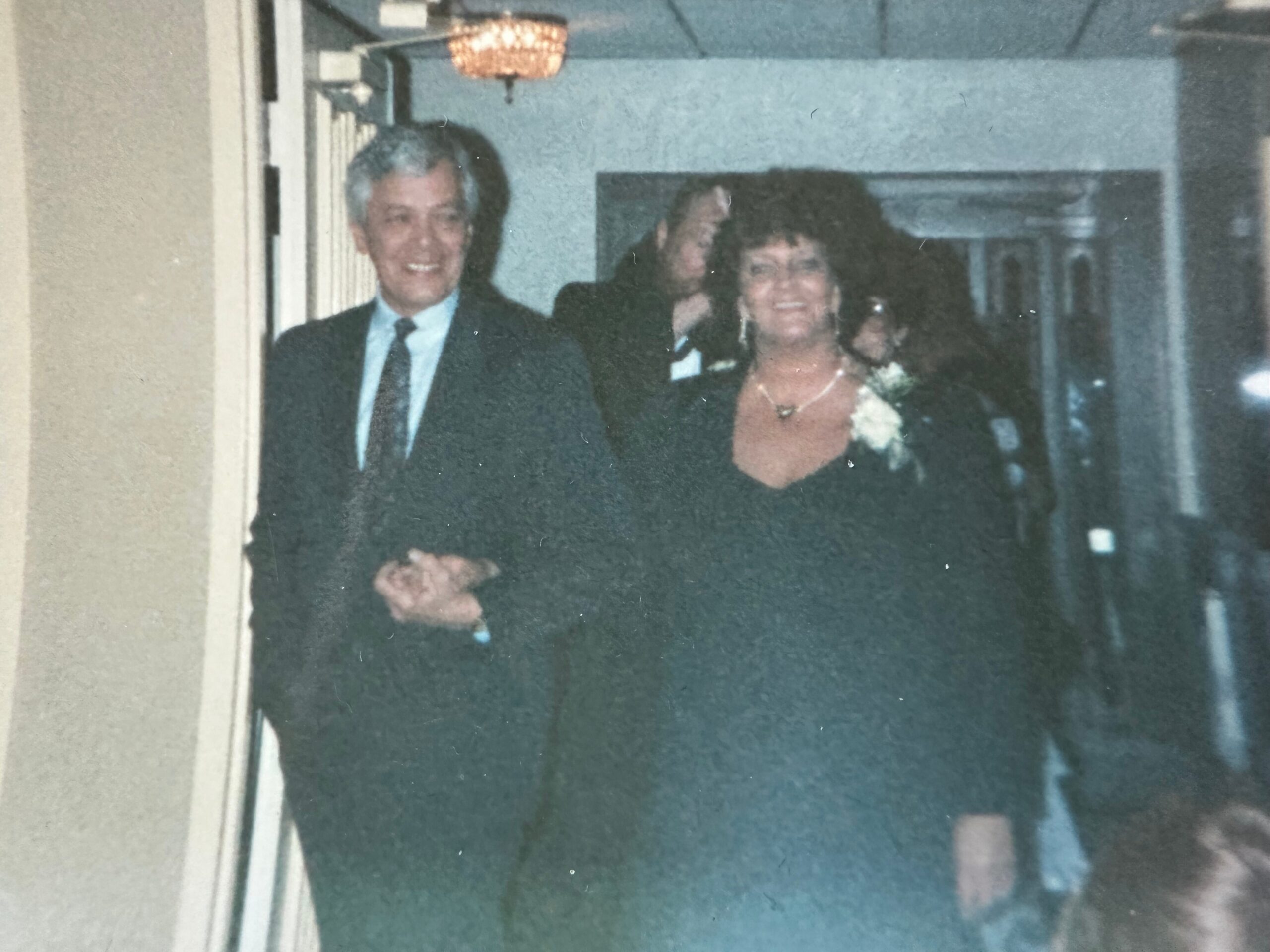 Smiling couple in formal attire walking down a dim hallway, a chandelier overhead.