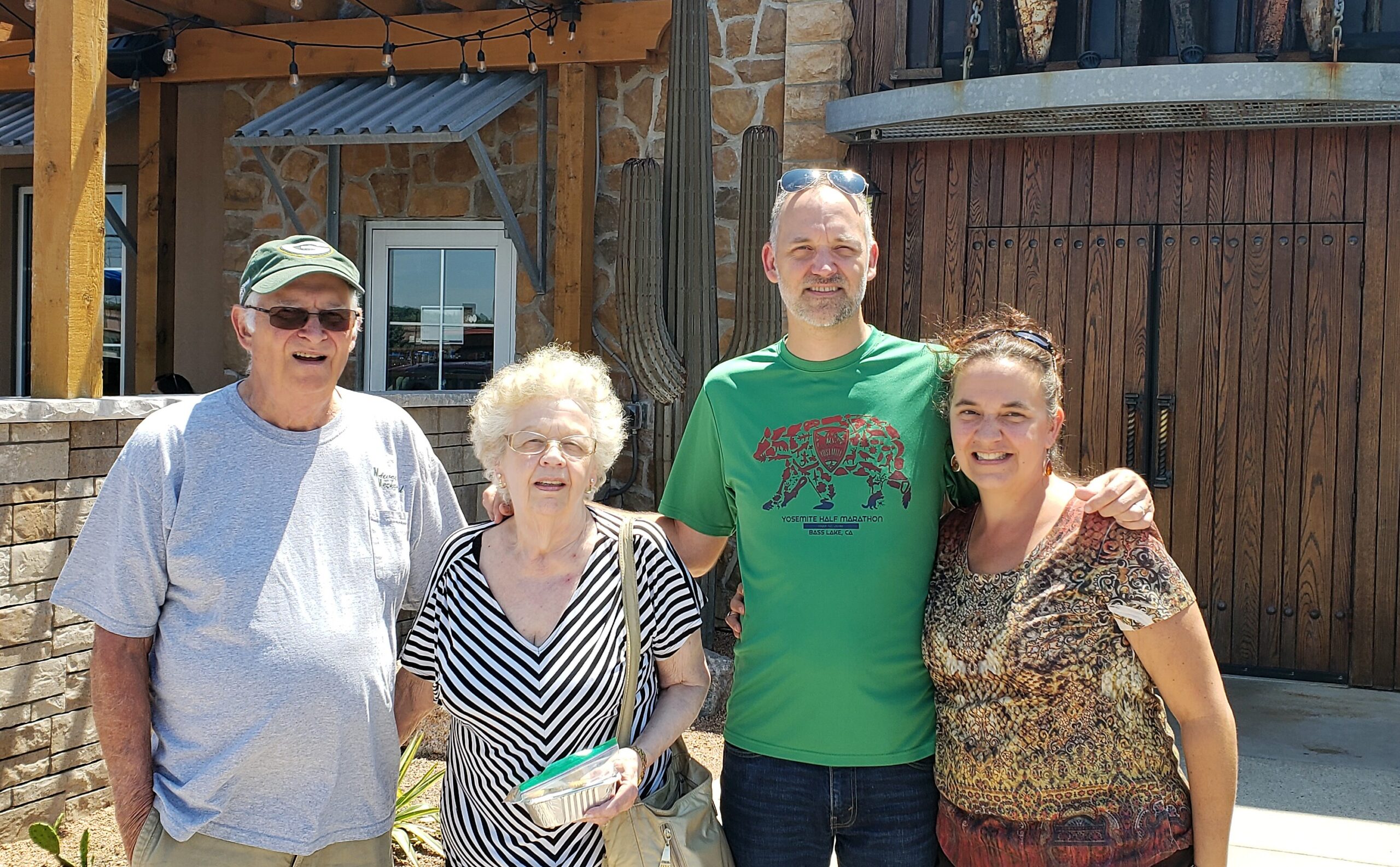 Four adults pose together outdoors in front of a stone and wood building on a sunny day, smiling for the photo.