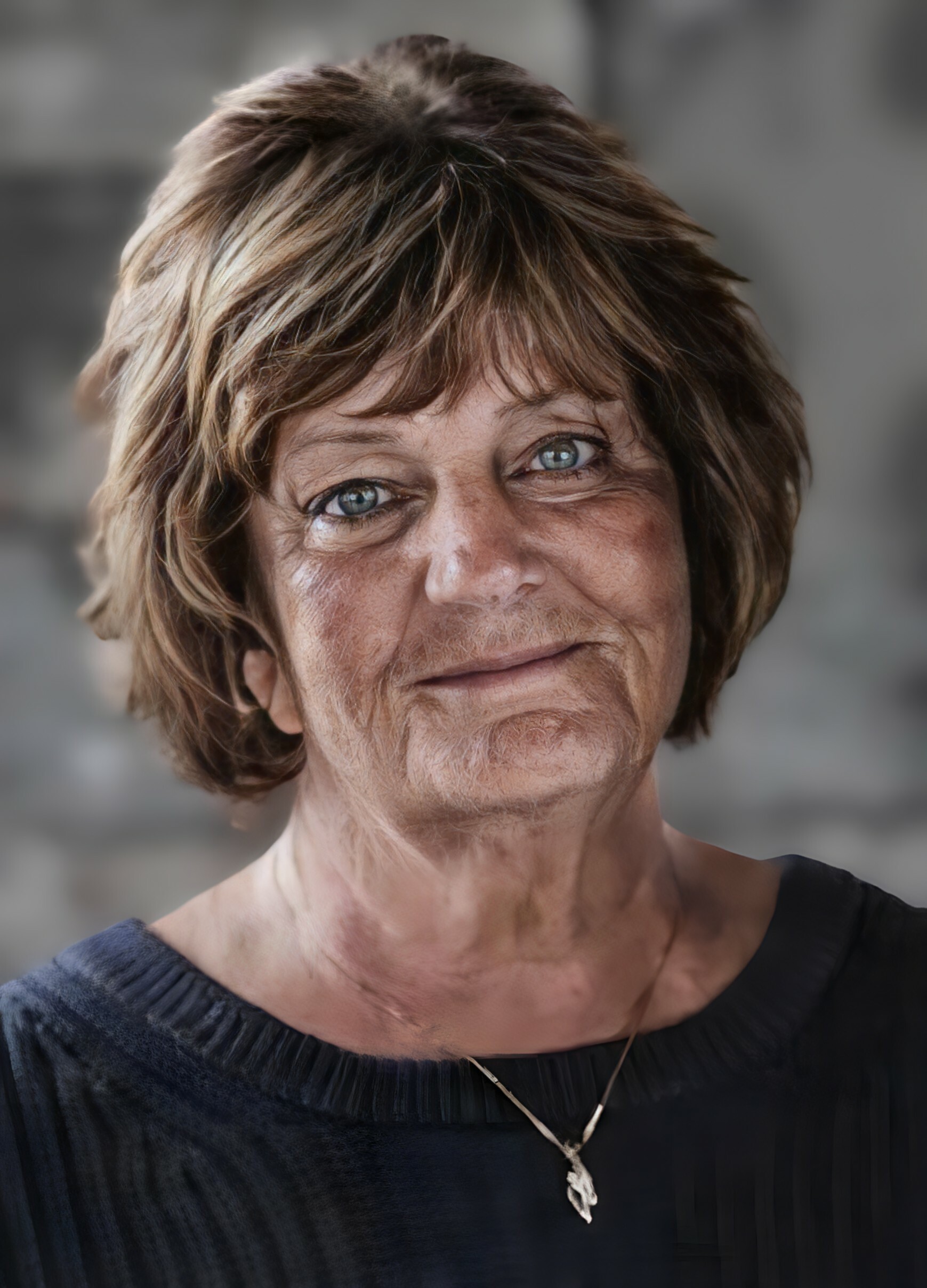 Close-up portrait of a smiling woman with short light brown hair and blue eyes, wearing a dark top and a pendant necklace.
