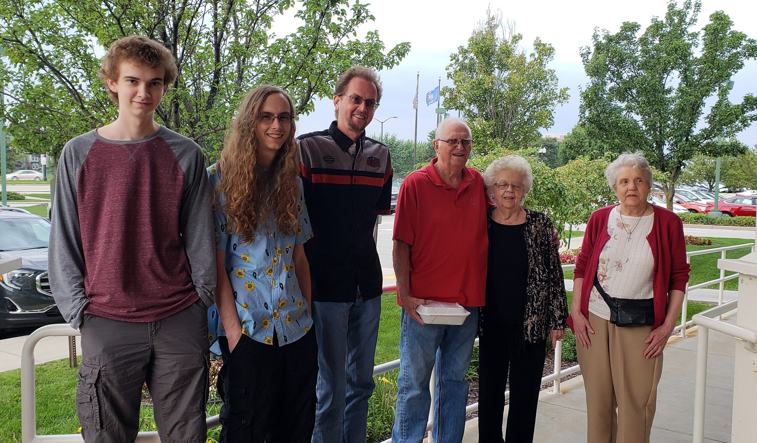 Six people of varying ages stand together outdoors on a sidewalk with green trees in the background, posing for a group photo.