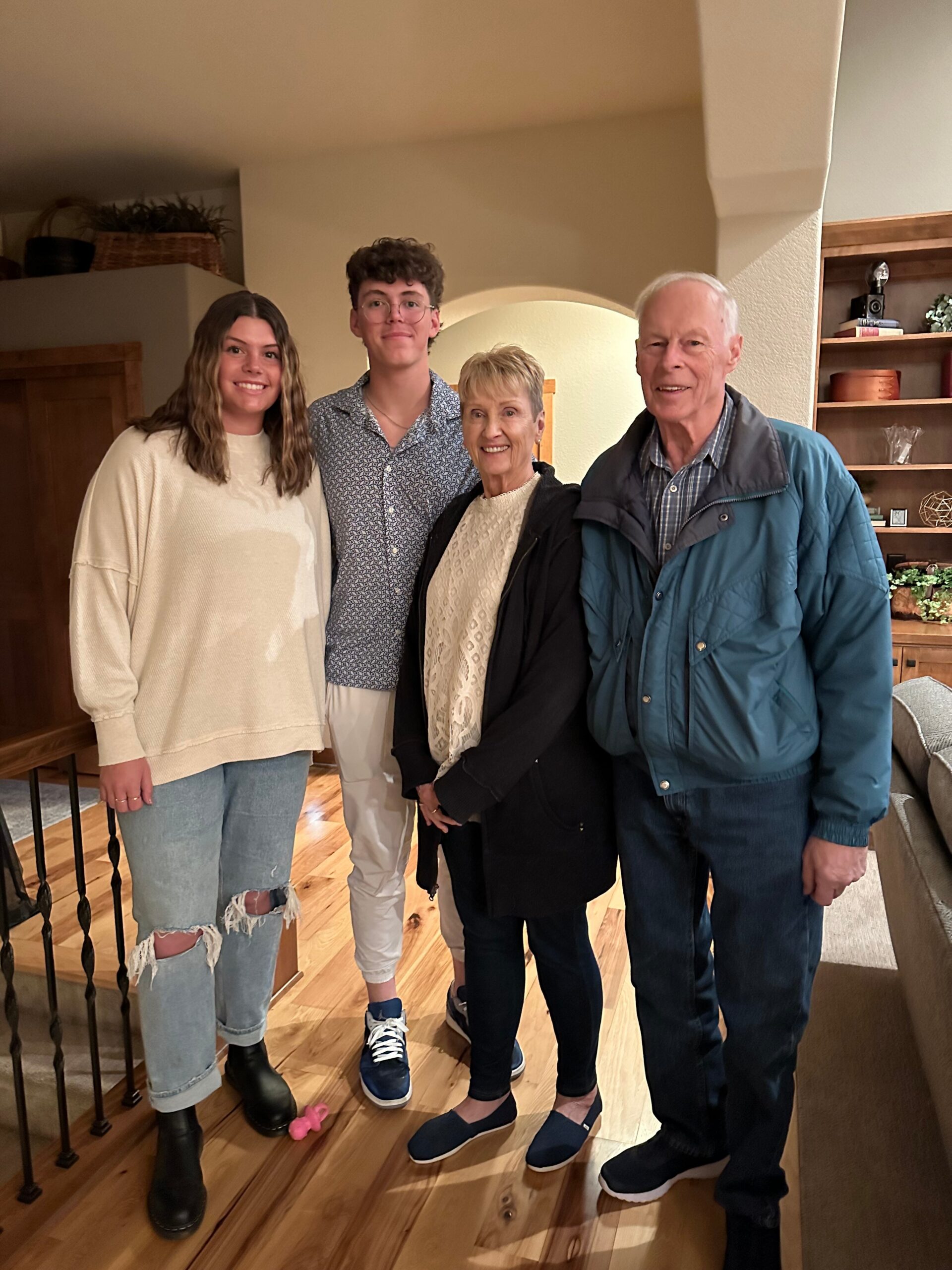 Four people indoors, smiling: a young woman in a beige sweater, a young man in a patterned shirt, an older woman in a cream sweater, and an older man in a teal jacket.