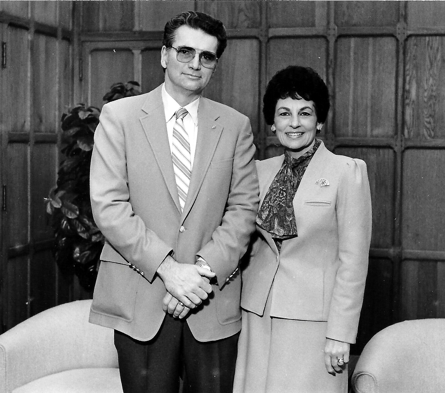 Man and woman in light-colored suits posing for a formal portrait indoors, both smiling.