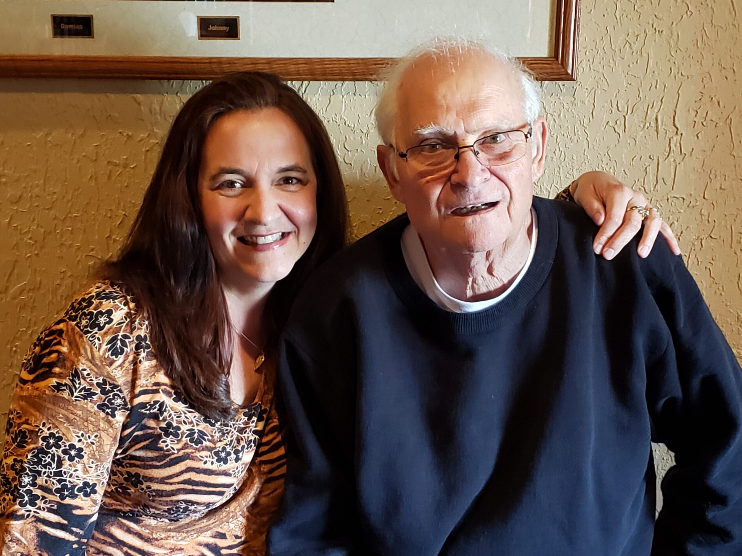 Two people smiling for a selfie, a woman on the left in a patterned top and an older man on the right with glasses, arm around each other.