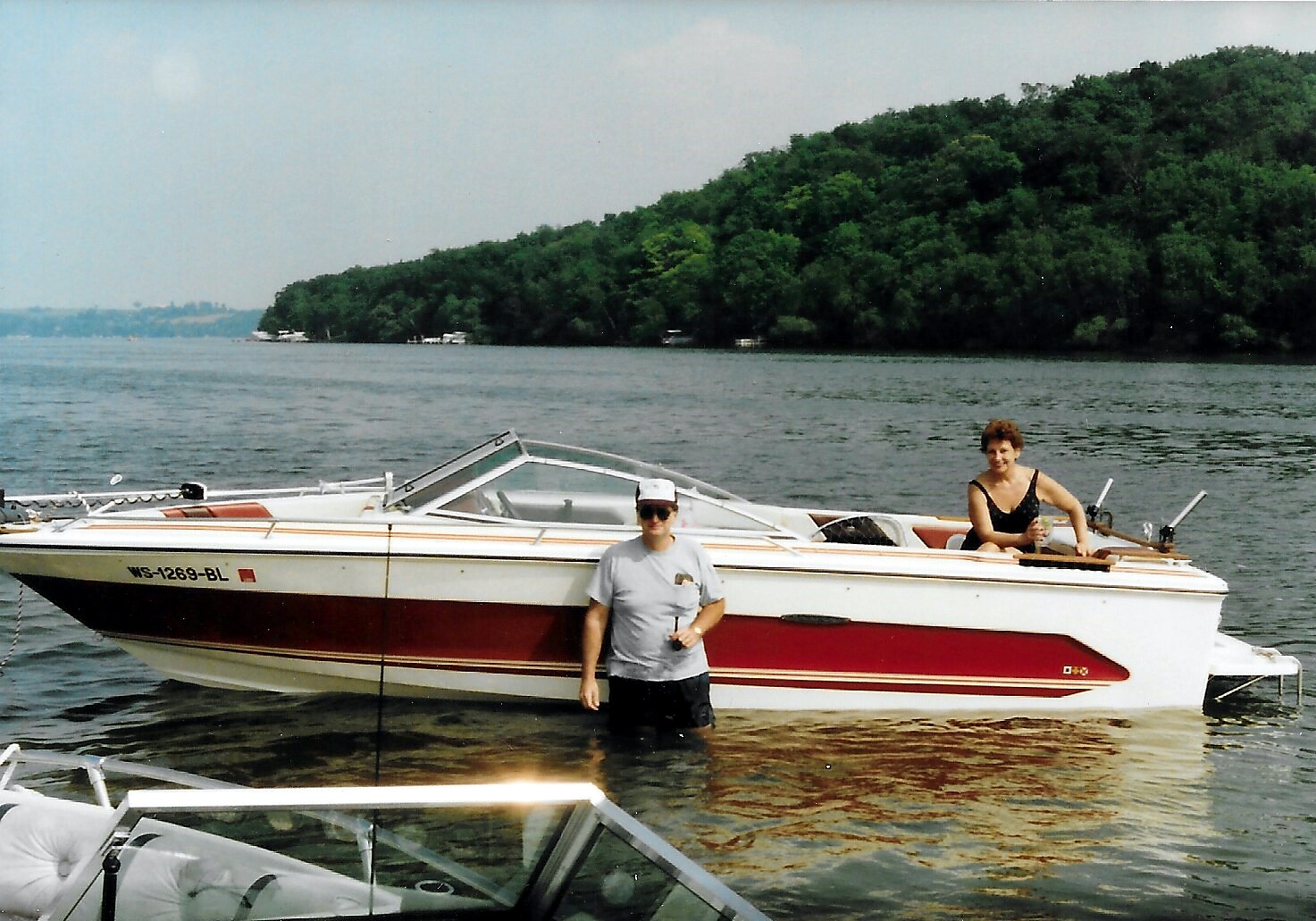 Two people on a lake with a red-and-white speedboat; one person stands in knee-deep water while the other sits in the boat near a tree-covered shoreline.