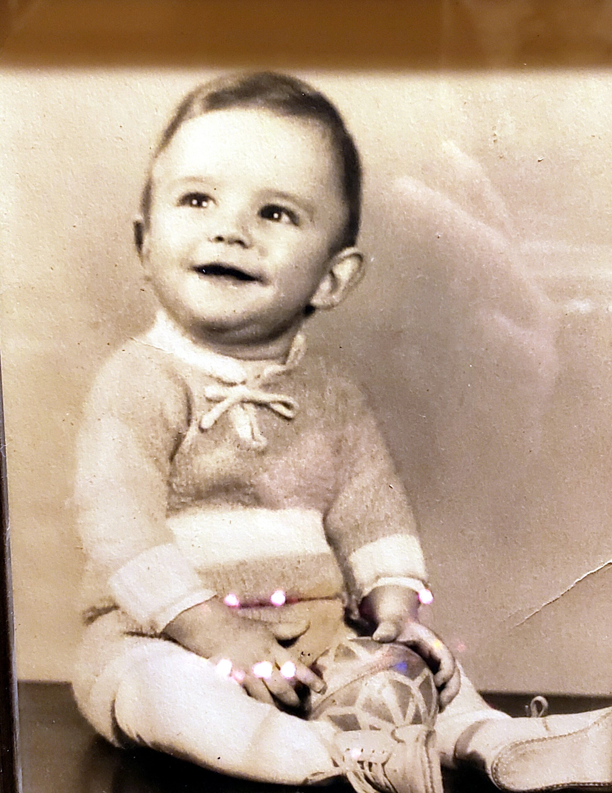 Smiling baby seated, wearing a knit sweater and bow, resting hands on a patterned ball in a vintage photo style.