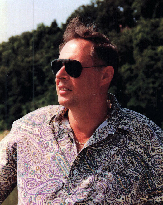 Man wearing dark sunglasses and a paisley shirt outdoors, looking to the left.