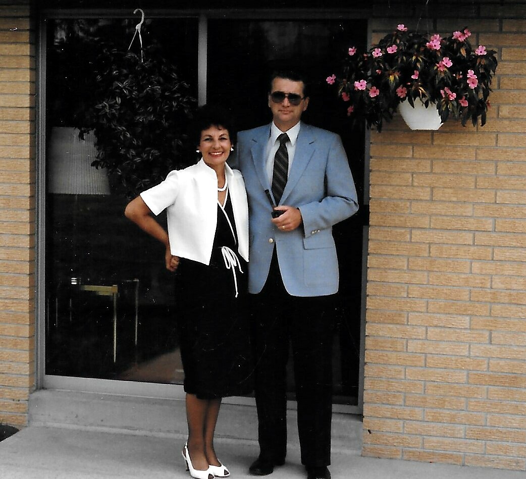 Smiling couple posing at a brick doorway; woman in a white blazer over a black dress, man in a light-blue suit with sunglasses.