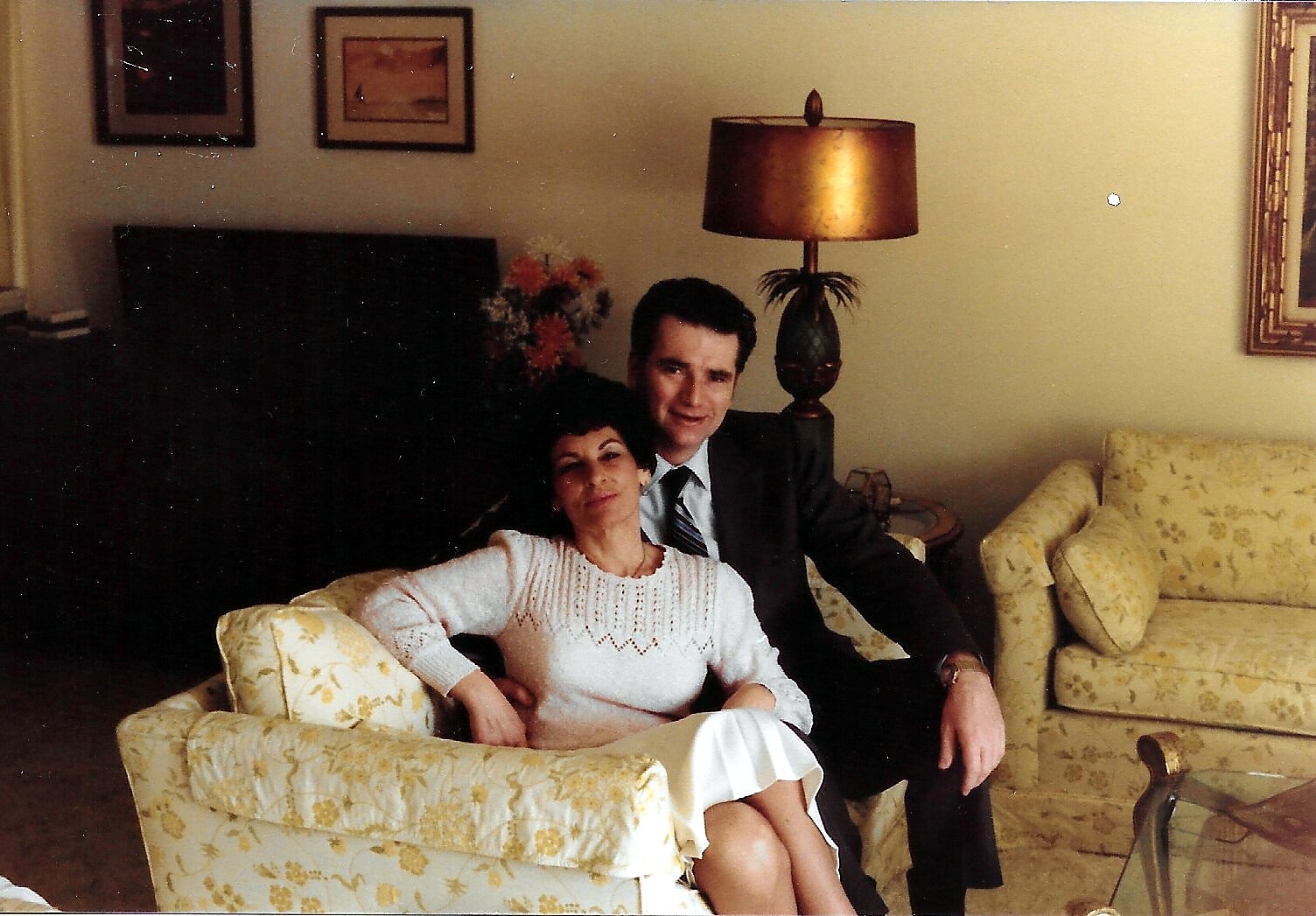 Couple relaxing on a floral yellow sofa in a living room, the woman leaning into the man as they smile at the camera.