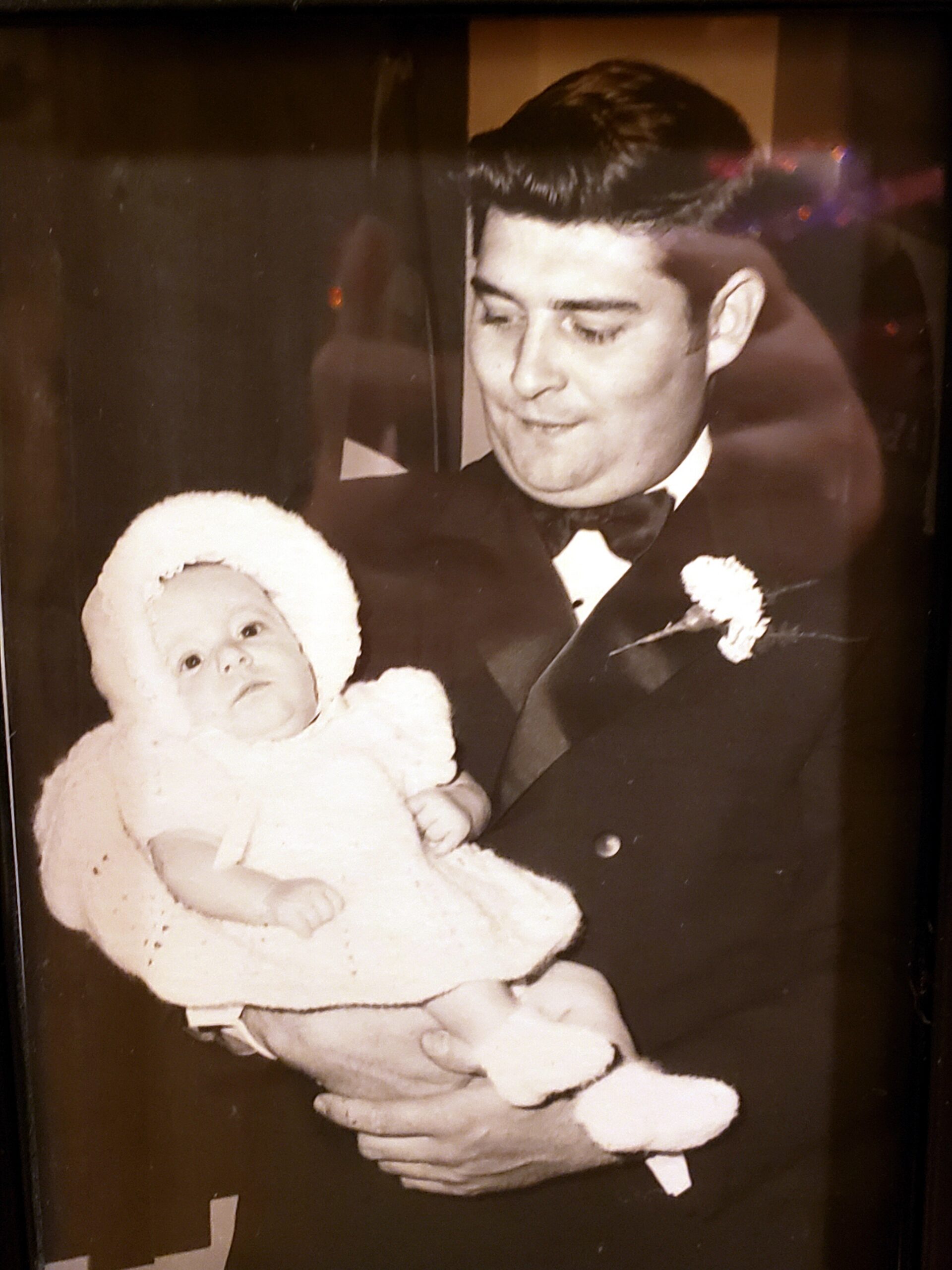 Man in a tuxedo gently holds a baby wrapped in a white, fluffy outfit for a formal photo.