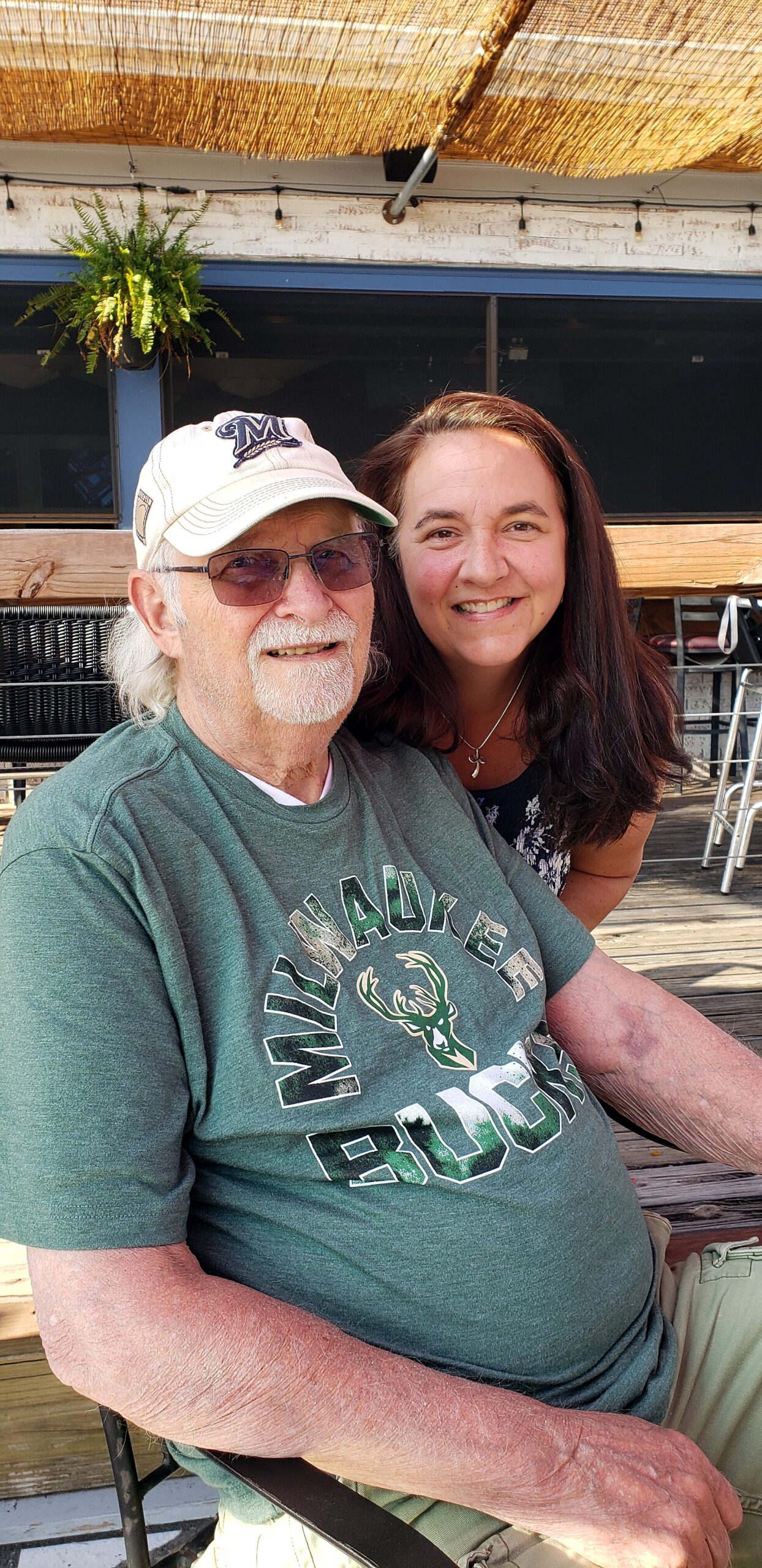 An elderly man wearing a white cap and sunglasses sits with a smiling woman beside him on a wooden outdoor deck.