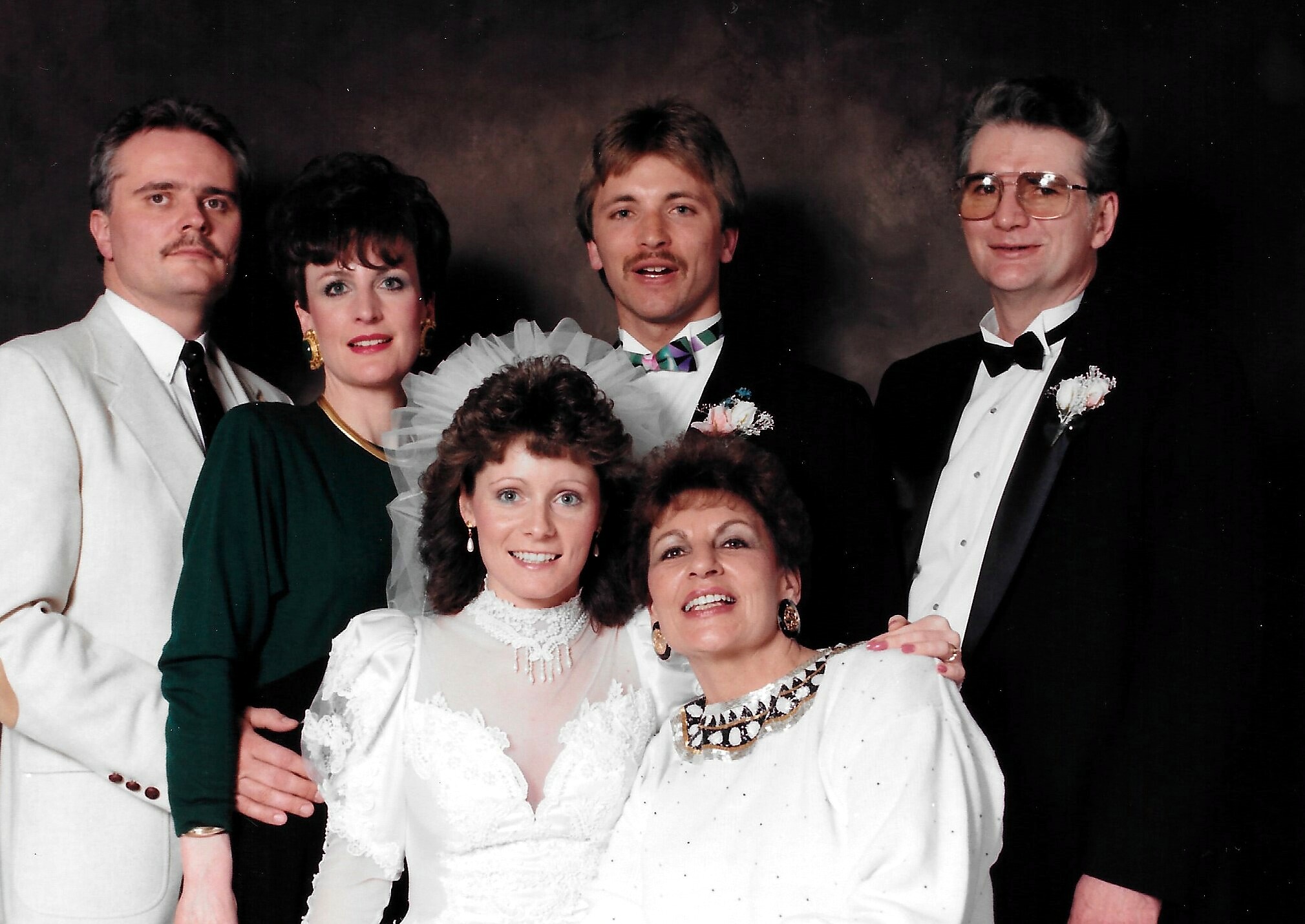 Wedding party photo: bride in white lace dress at center with guests in formal attire around her, smiling.