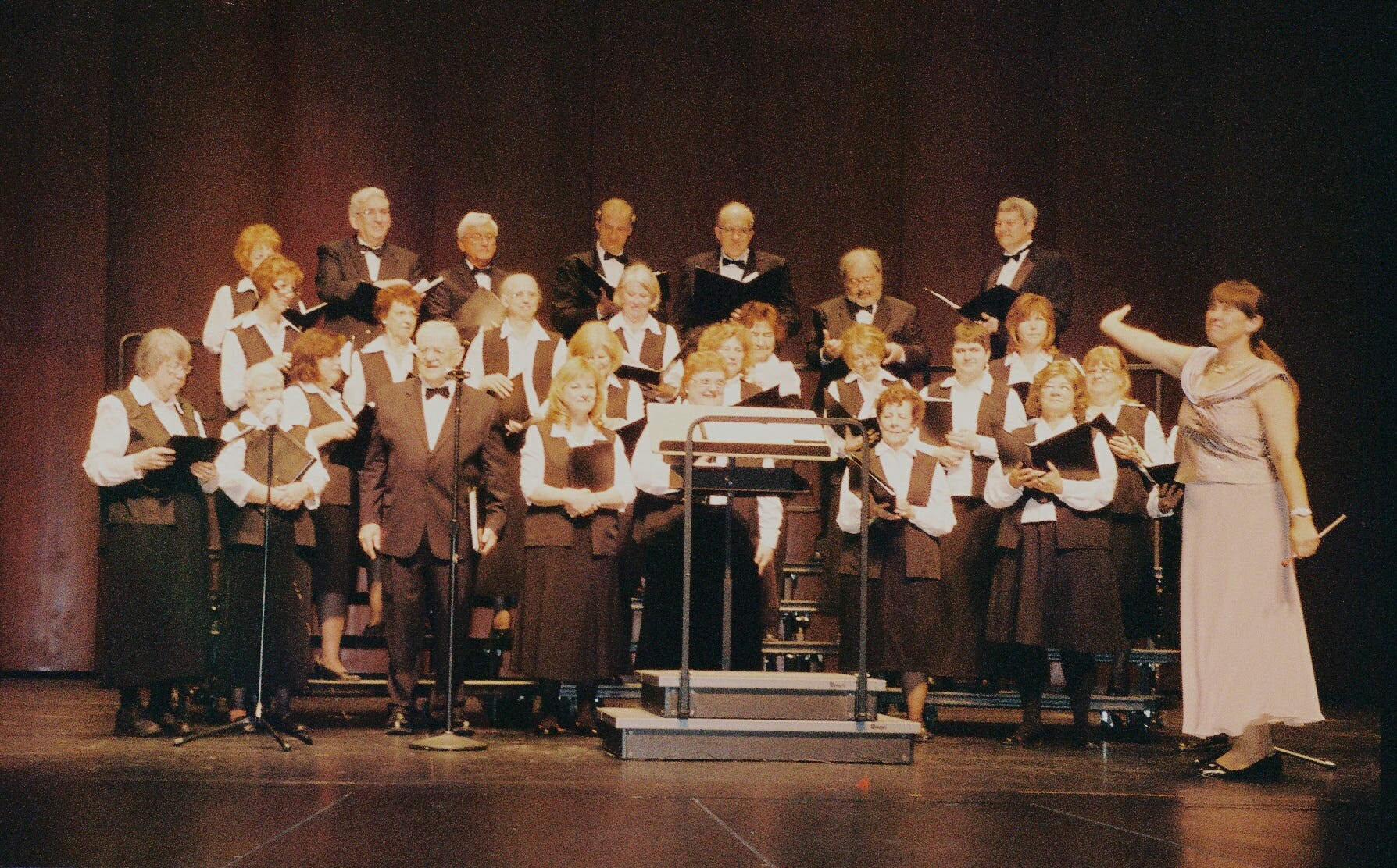 Large choir of men and women in formal black-and-white attire on stage, with a female conductor in a light dress directing from the right.