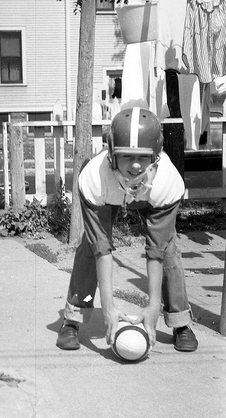 Boy in a football helmet and striped shirt crouches over a ball on a concrete path in a suburban yard, ready to snap.