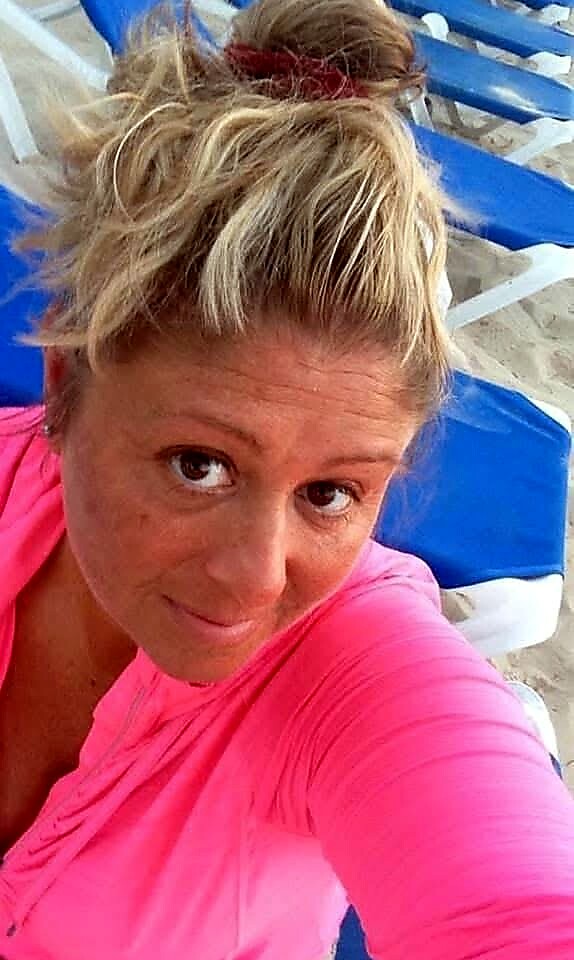 Woman with a messy blond bun wearing a bright pink top on a sunny beach, blue lounge chairs in the background.