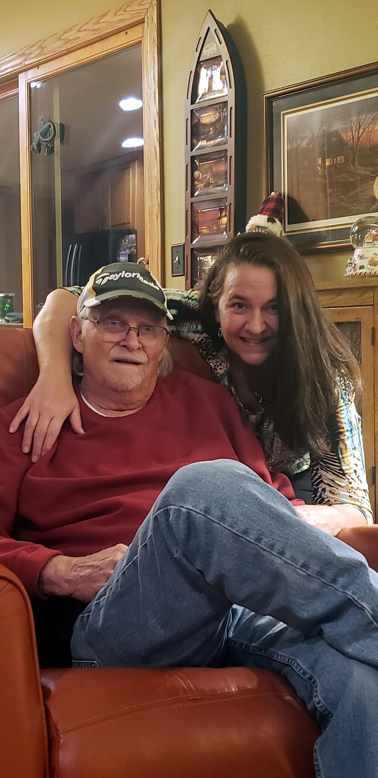 An older man in a red sweatshirt and glasses sits on a brown leather chair while a woman hugs him from the side, smiling in a cozy living room with wood paneling and framed art behind them.