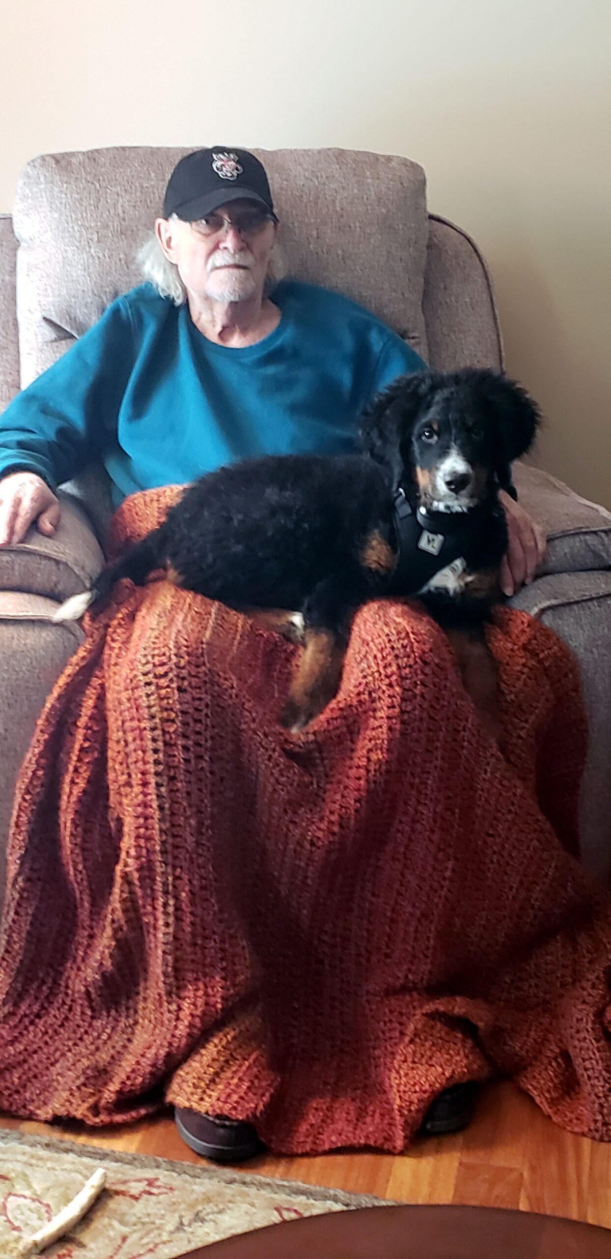 Older man wearing a teal shirt and black cap sits in a beige chair, with a small black and tan dog on his lap, covered by an orange blanket.