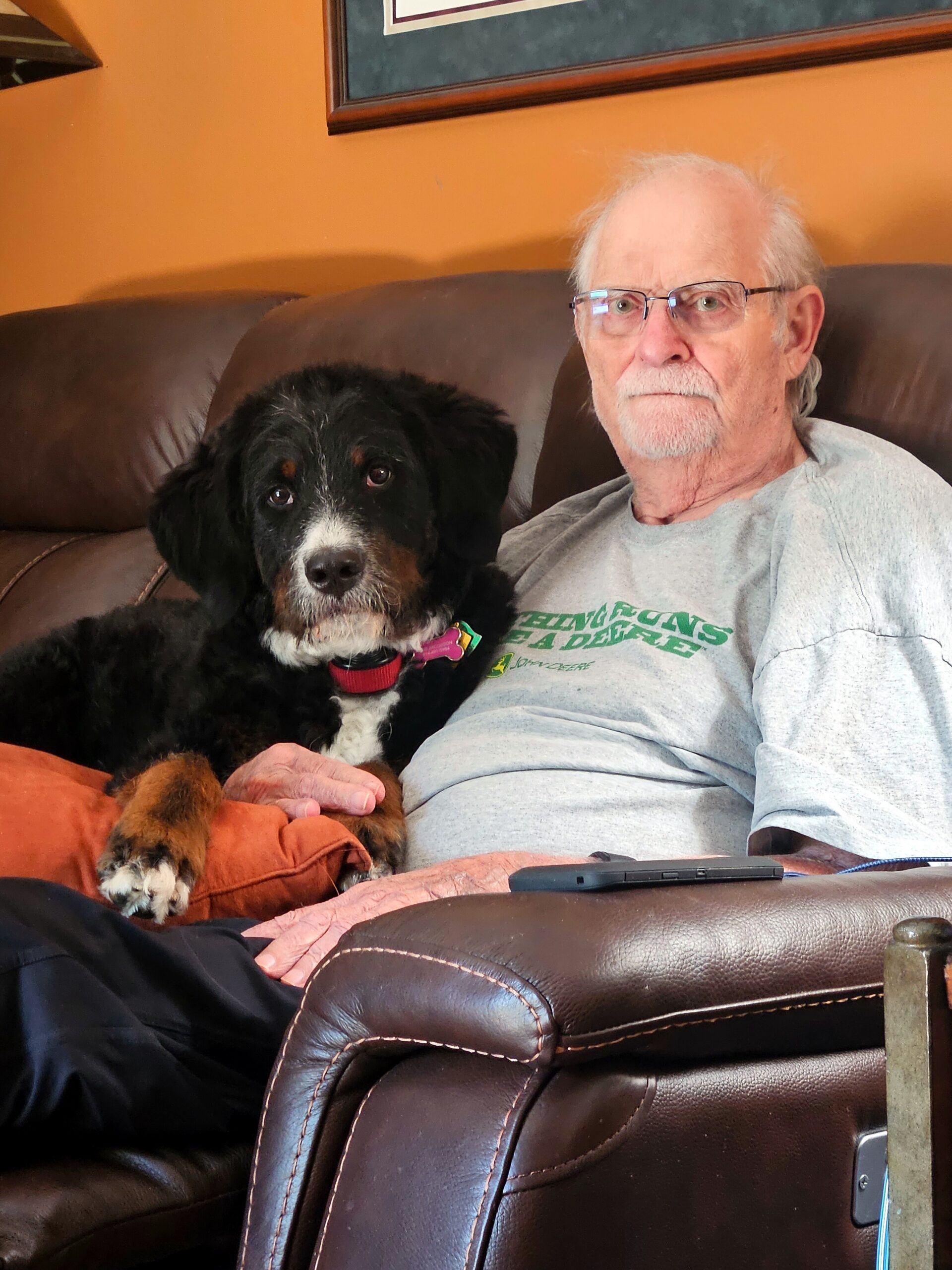Older man with glasses relaxing on a brown leather sofa with a black and white dog cuddled beside him, wearing a gray shirt.