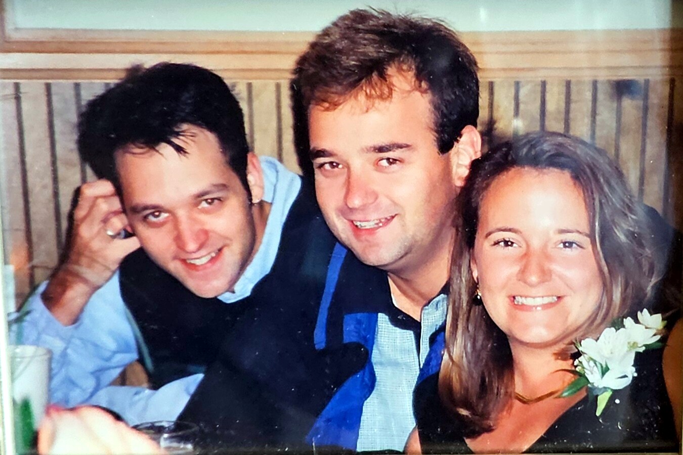 Three friends posing closely for a photo at a celebration: two men on the left and center, and a smiling woman on the right wearing a white corsage eyeing the camera.
